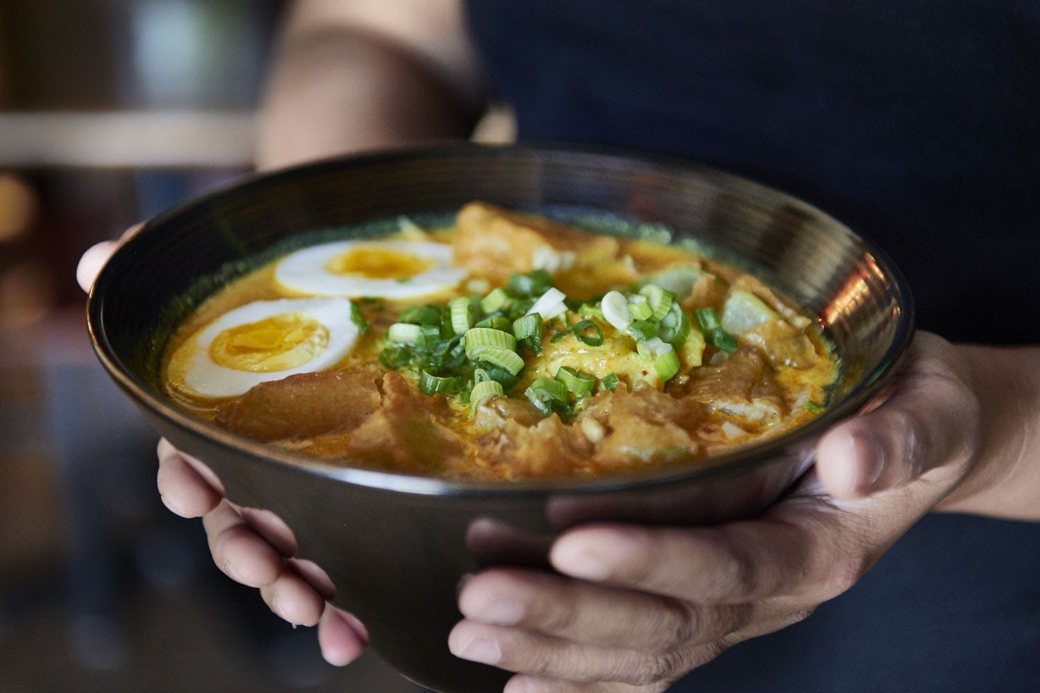 Hands holding a bowl of ramen topped with sliced egg and green onions