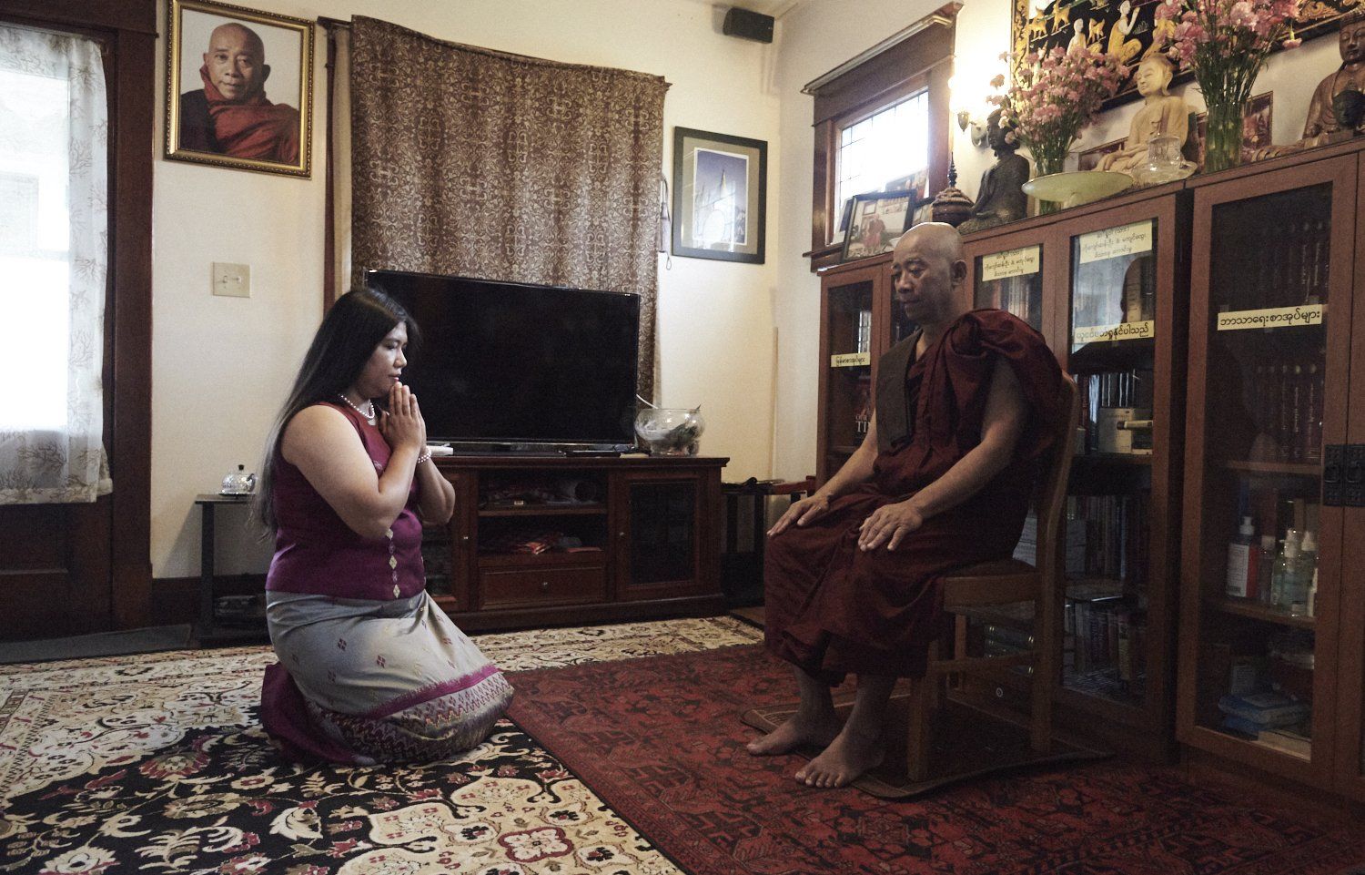 Two people kneel facing each other in a living room with rugs, curtains, and a TV.