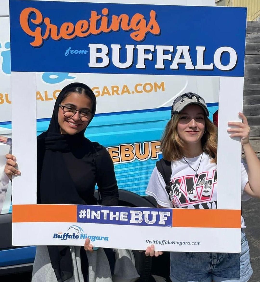 Two people pose at a “Greetings from Buffalo” sign, one holding the frame.