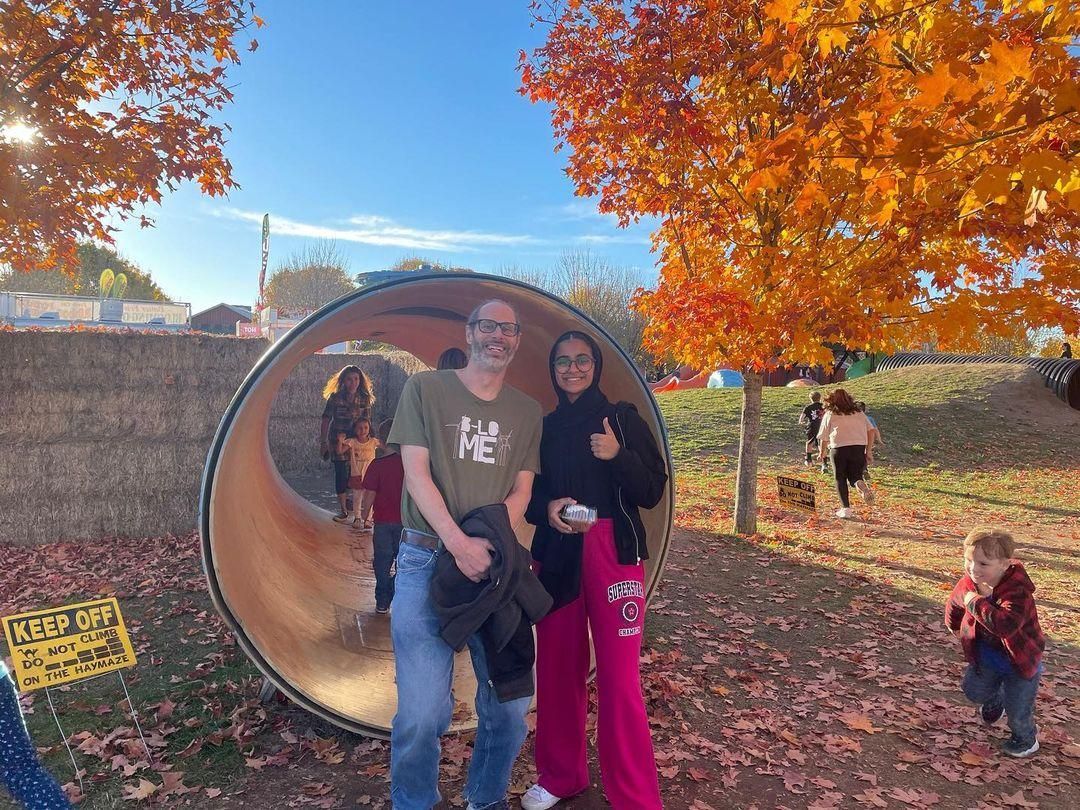 Two people posing by a large tunnel at a fall festival with orange leaves and pumpkins nearby