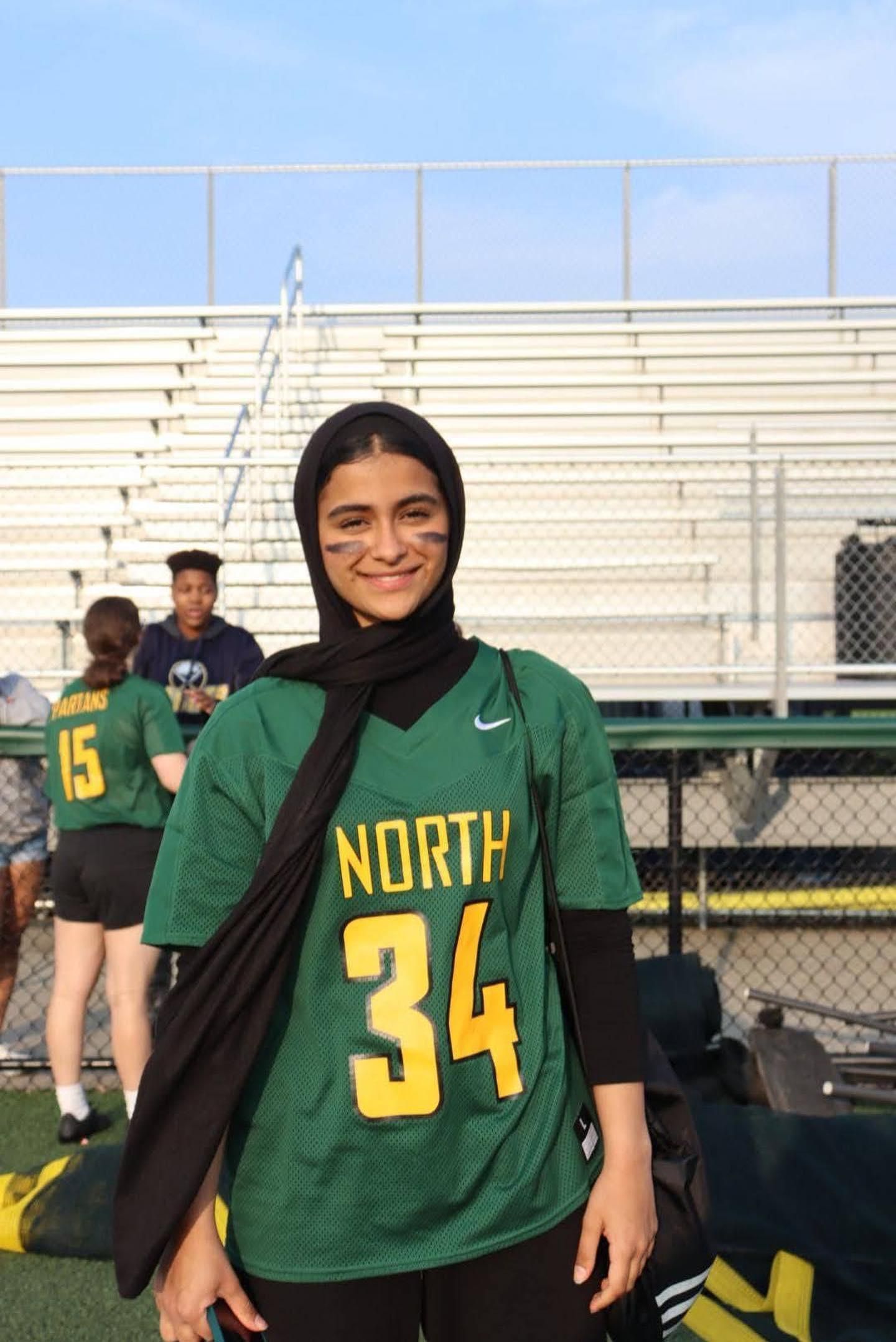 Person in green North jersey and black scarf standing on a sports field, smiling in front of bleachers.