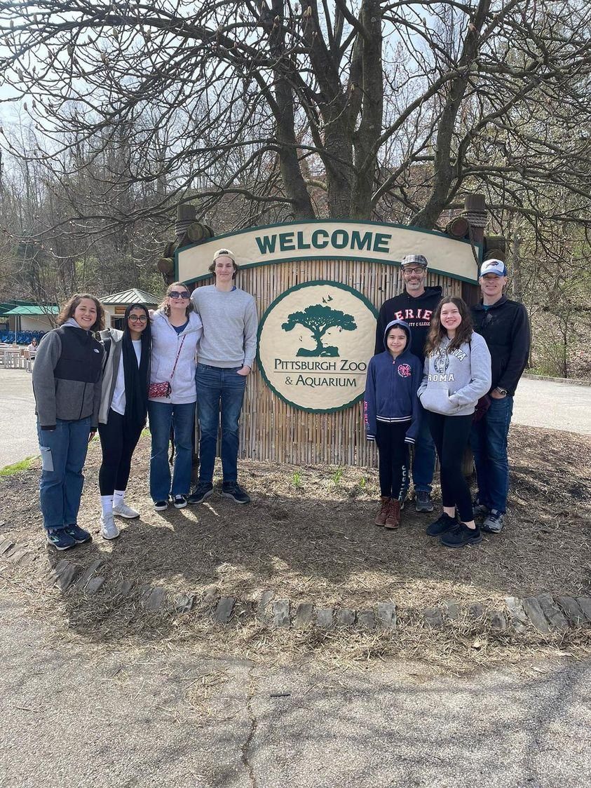 Group posing by a Welcome sign in a wooded outdoor area on a sunny day