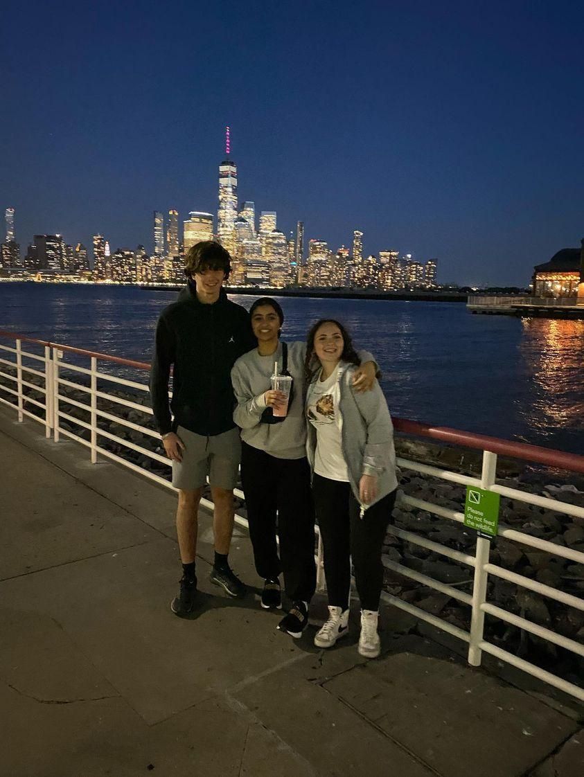 Three people posing by a waterfront railing at night with a lit city skyline across the water.