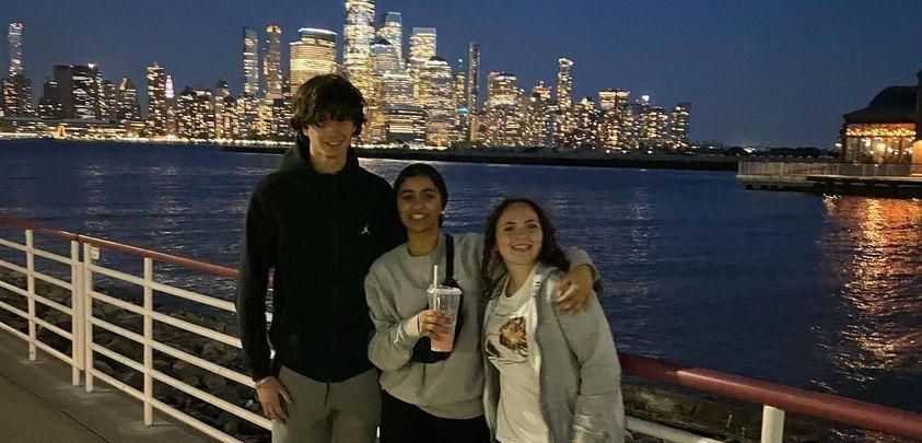Three people pose by the water at night with a lit city skyline in the background.