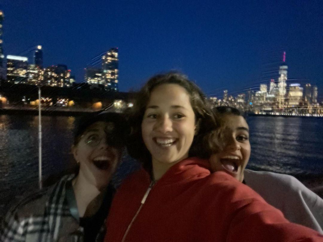 Three smiling friends taking a selfie at night by the water, with city lights in the background.