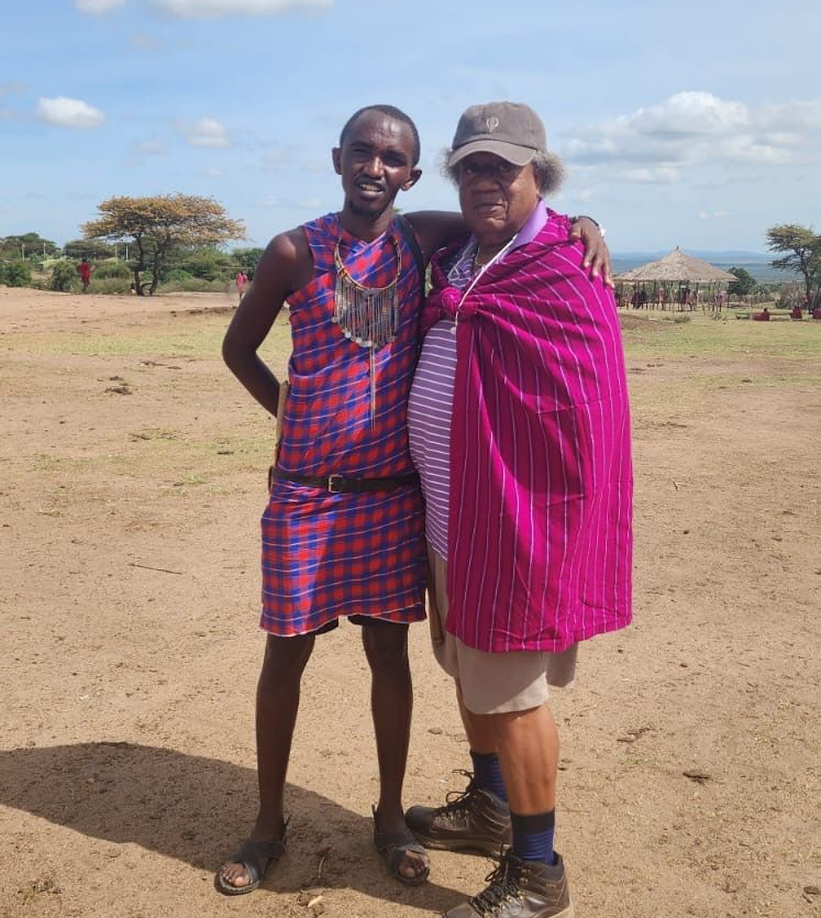 Two people standing together outdoors on a dirt field, smiling in colorful clothing under a blue sky.