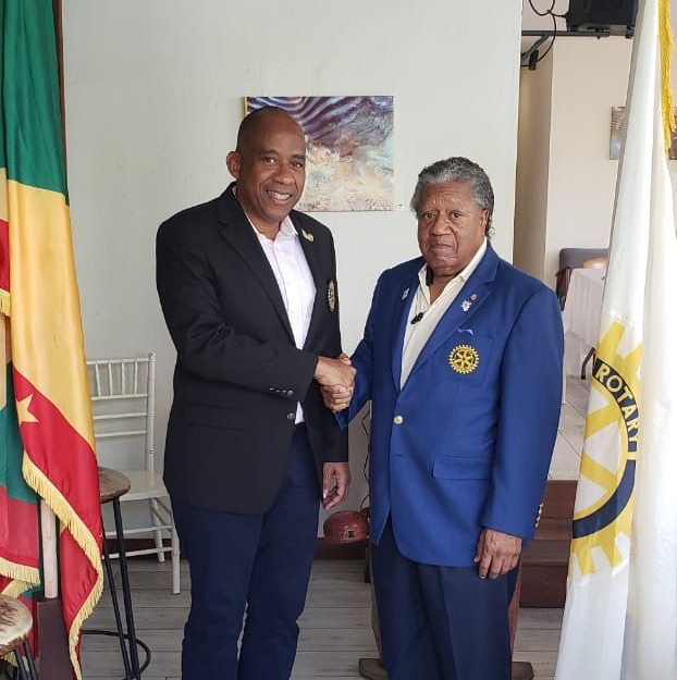 Two men in suits shaking hands indoors beside flags and a framed painting