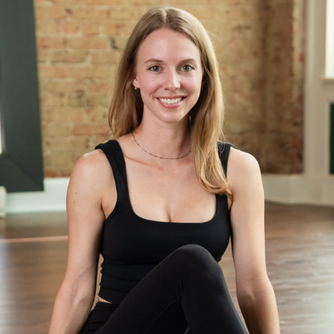 Amy Ankli, an Uptown Yoga instructor in Dallas, smiling while sitting on a wood floor in a studio wearing black activewear.