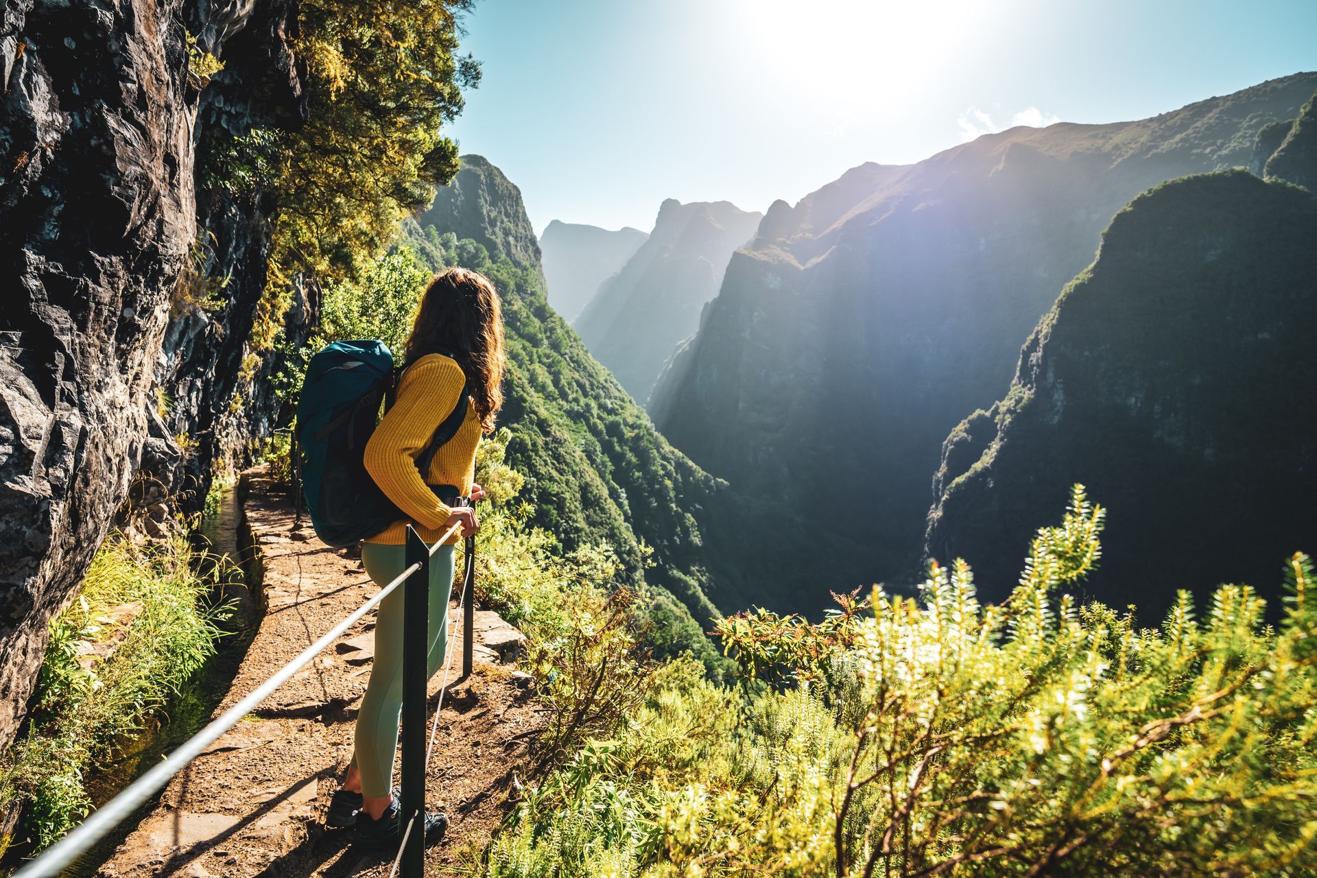 Woman with a backpack hiking a scenic cliffside trail overlooking a sunlit valley on a Madeira yoga retreat.