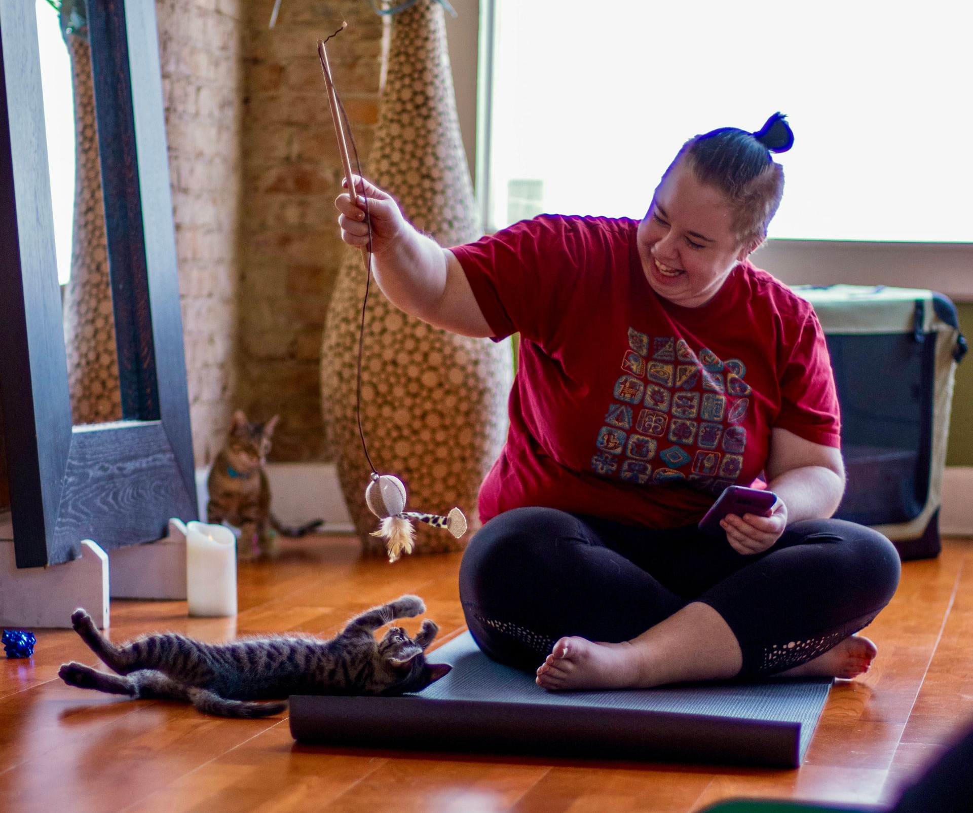 An Uptown Yoga student laughing and playing with a foster kitten rolling on its back using a feather wand toy during a donation-based event for Spicy Whiskers Rescue.