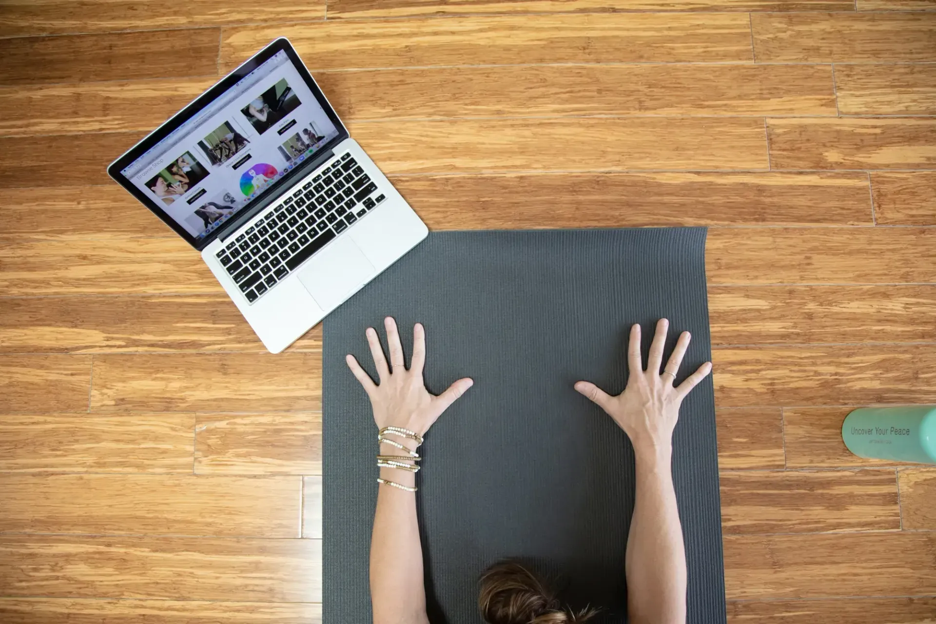 Top-down view of a woman with her hands on a yoga mat preparing for an online class on her laptop, demonstrating a home yoga practice setup.