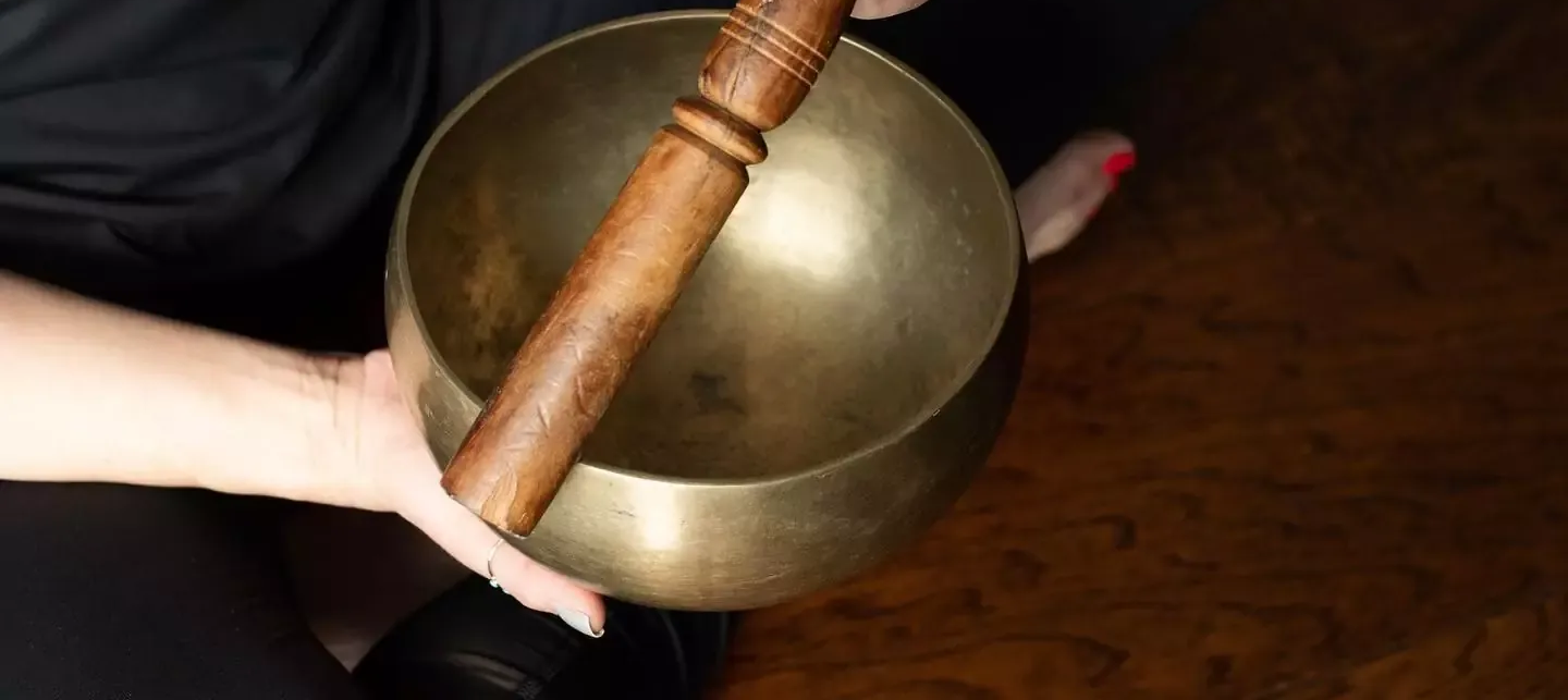 Hands holding a brass singing bowl and wooden mallet for a sound bath meditation and healing therapy session at Uptown Yoga.