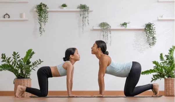 A woman and a young girl practicing cow pose together on a yoga mat in a bright, plant-filled room.