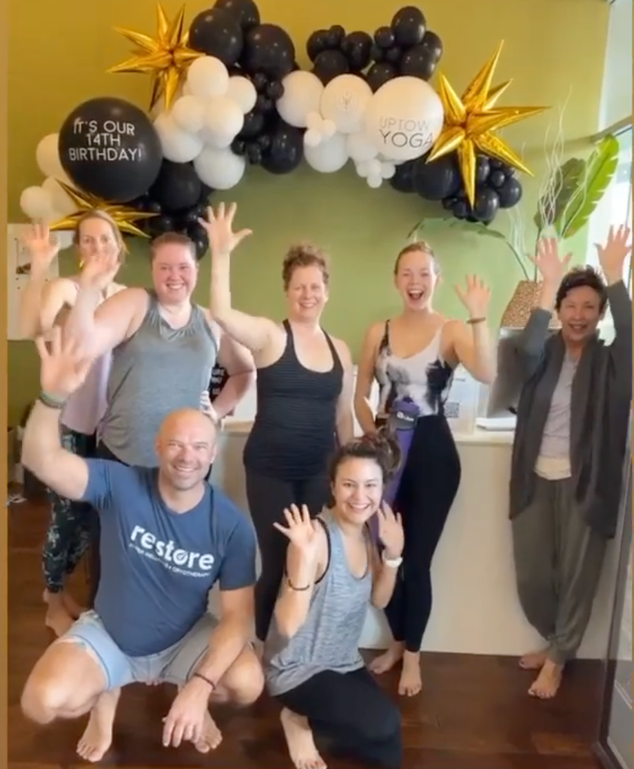Group of yoga instructors and students waving under a black and white 14th birthday balloon arch at Uptown Yoga.