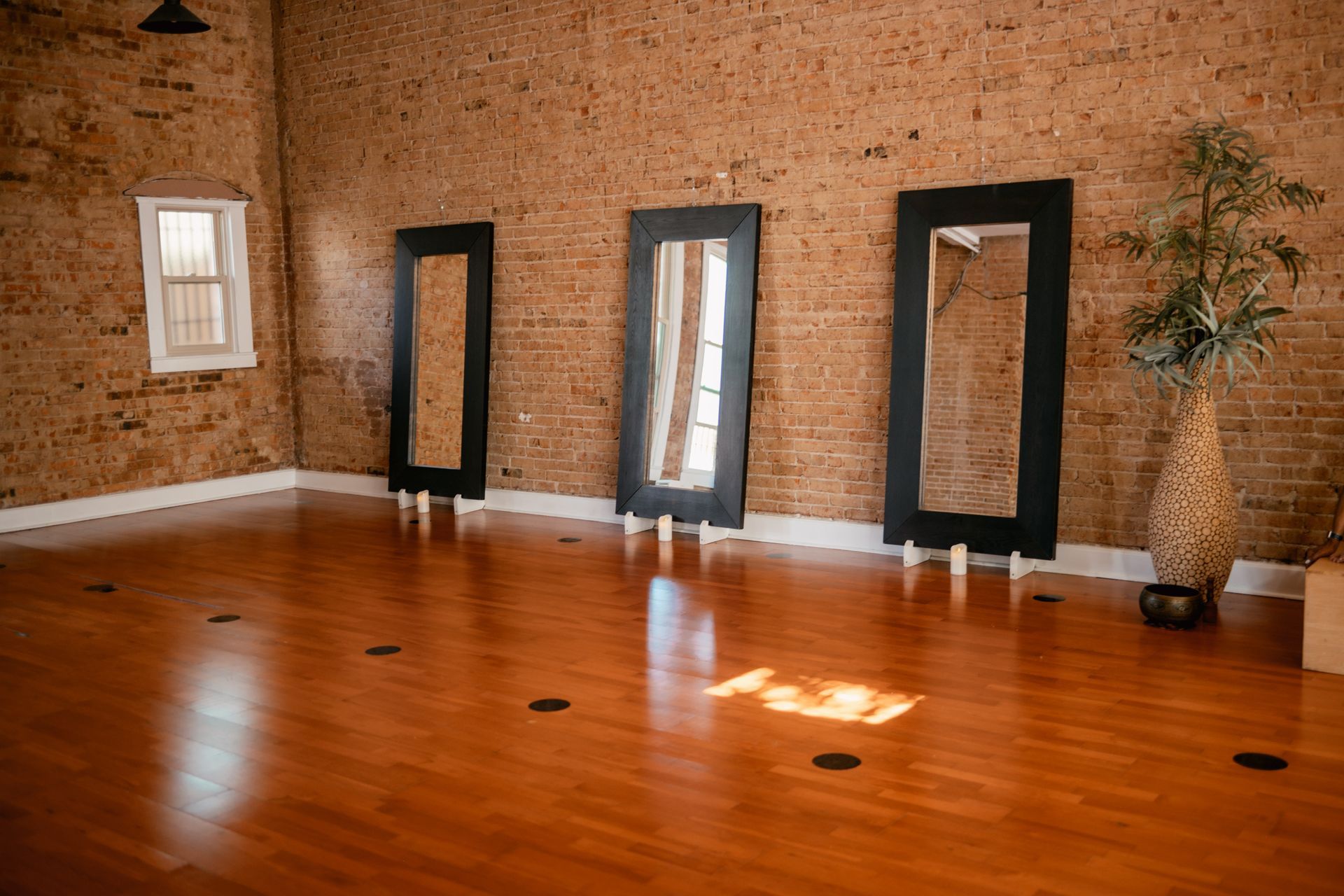 The spacious main yoga studio at Uptown Yoga in Dallas, featuring historic exposed brick walls, warm hardwood floors, and large leaning mirrors.