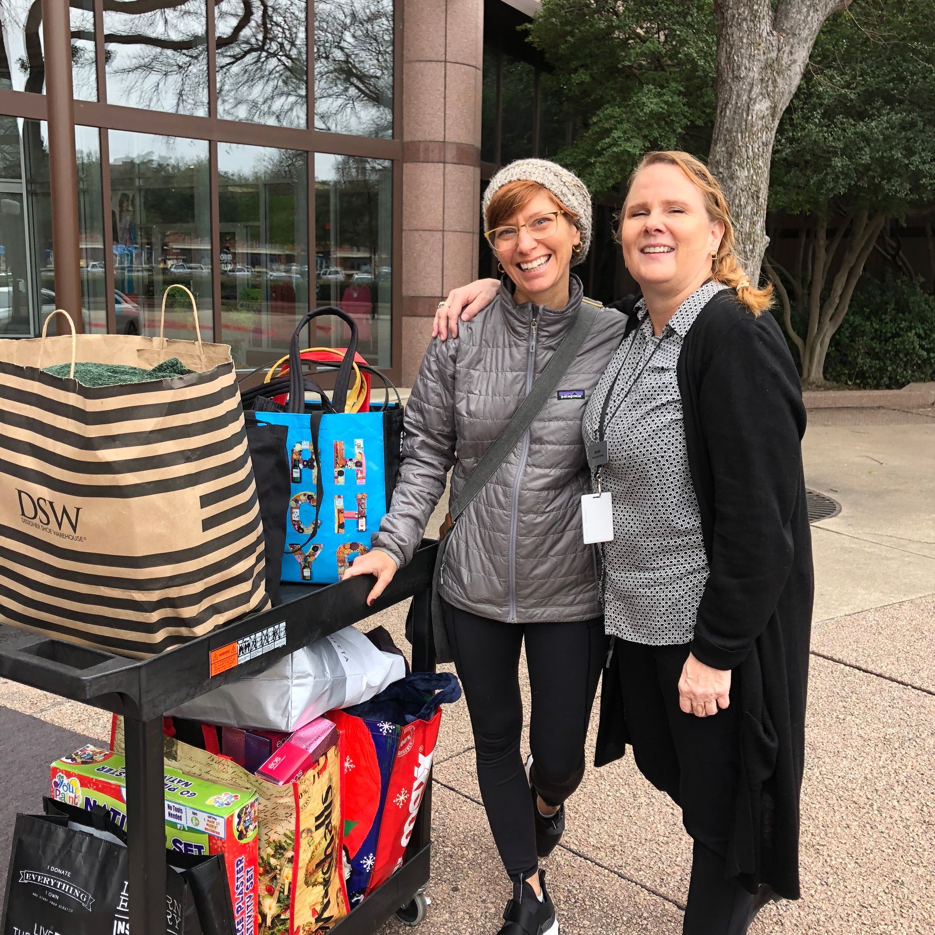 Two Uptown Yoga community members smiling next to a cart full of donated gifts for the Holiday Helpers drive supporting Mosaic Family Services.