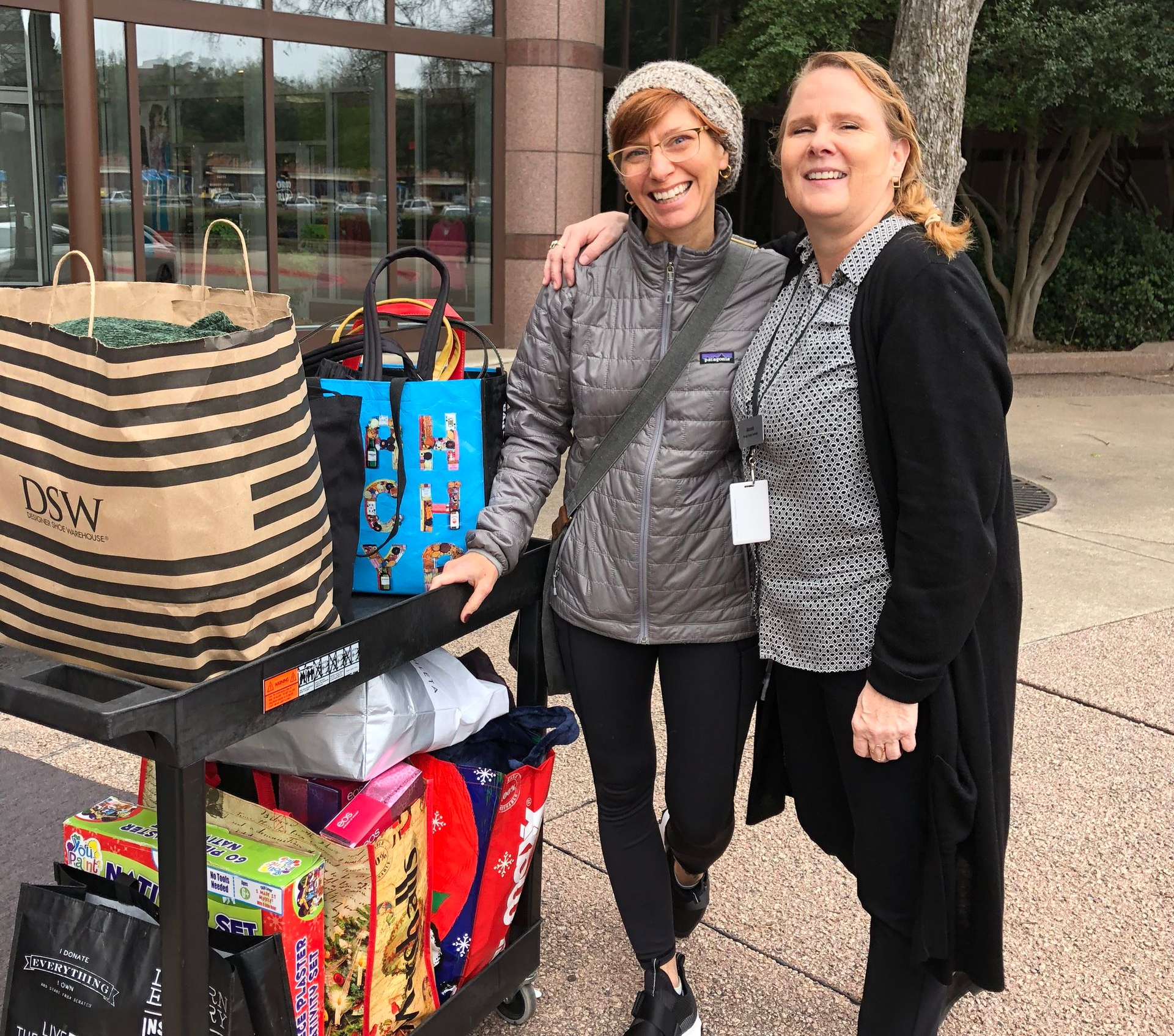 Two Uptown Yoga community members smiling next to a cart full of donated gifts for the Holiday Helpers drive supporting Mosaic Family Services.