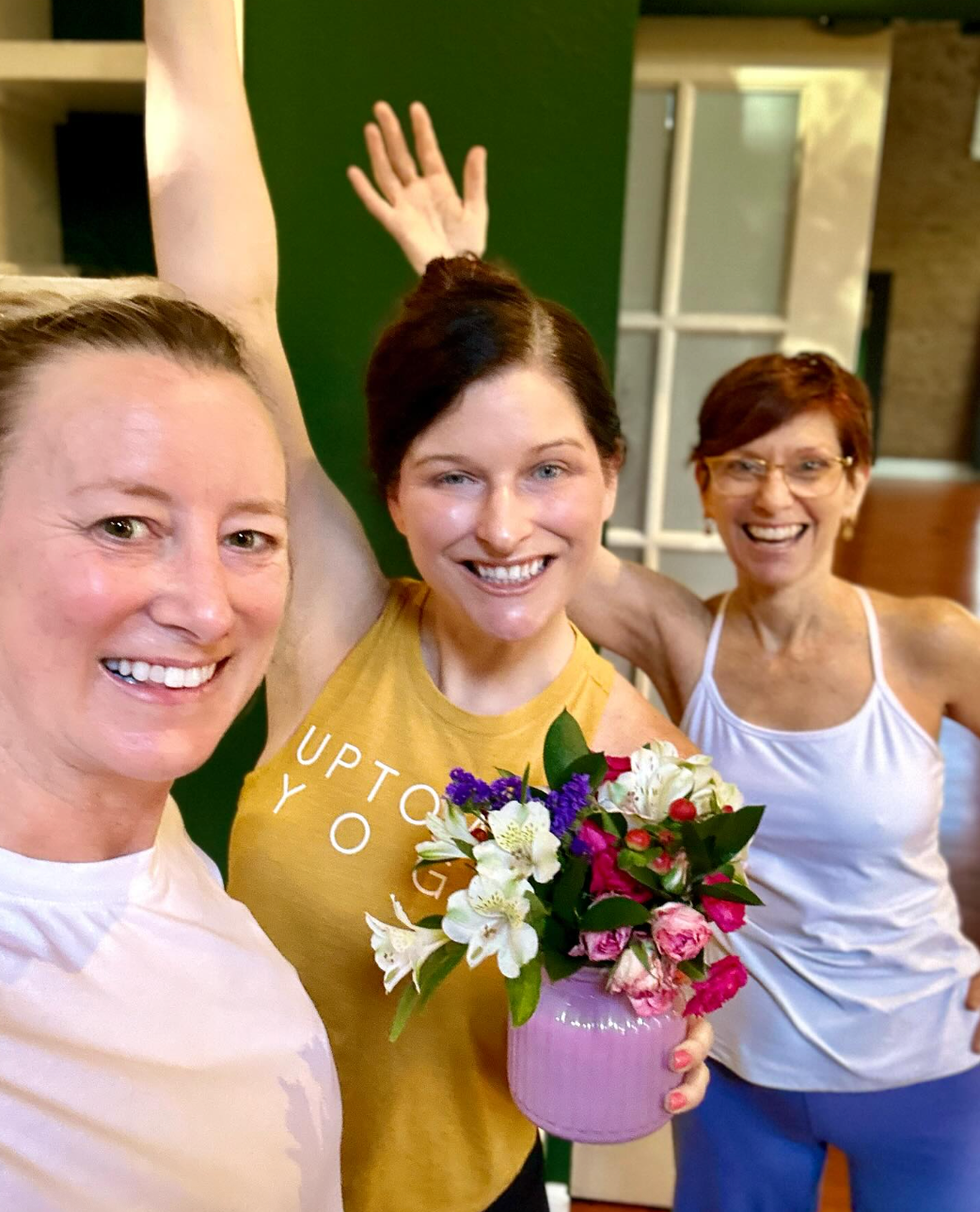 Three smiling women, one wearing a yellow Uptown Yoga tank top and holding a small purple vase with fresh flowers.