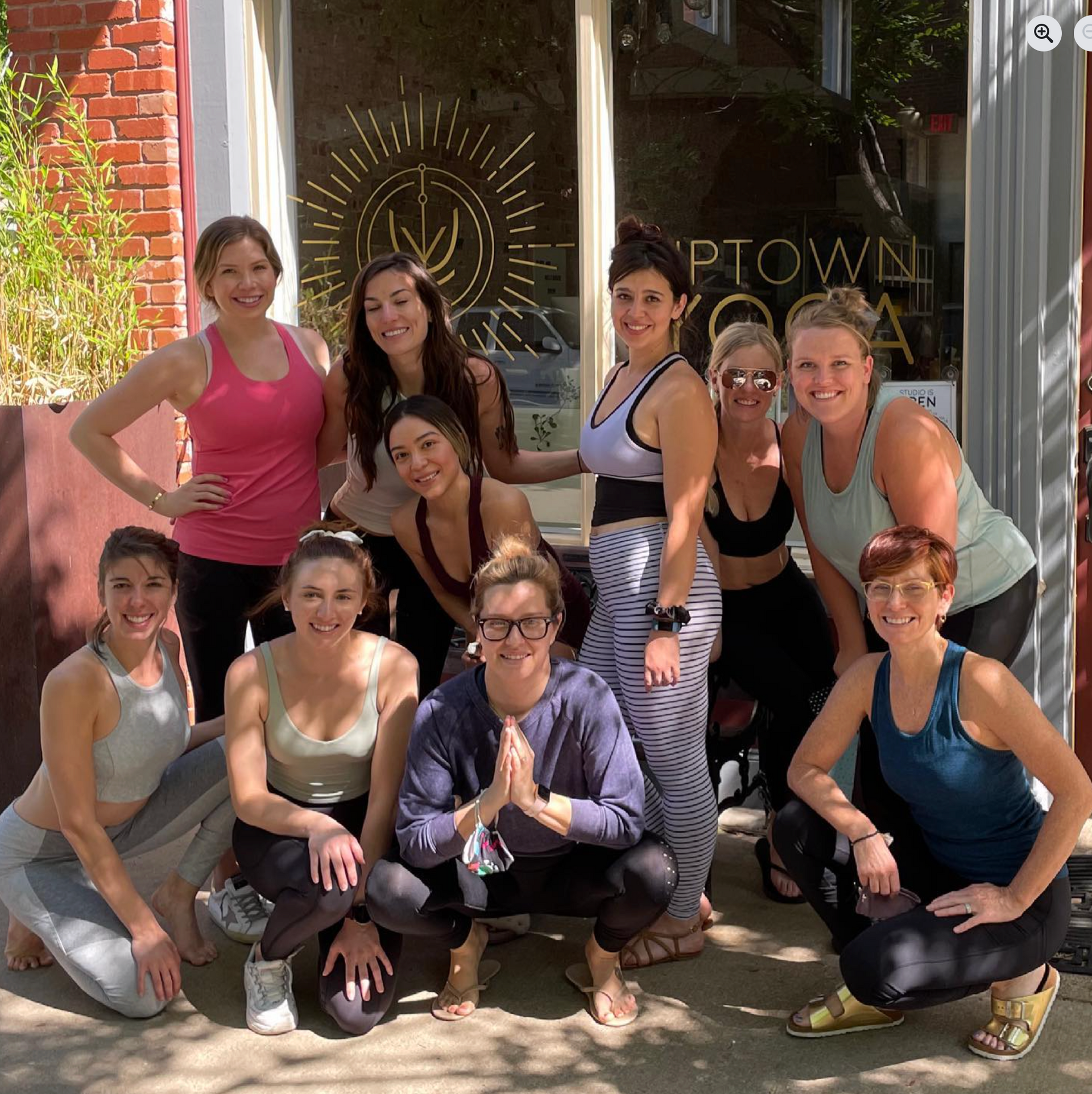 Group of smiling women in athletic wear gathered on the sidewalk outside the Uptown Yoga studio storefront window.