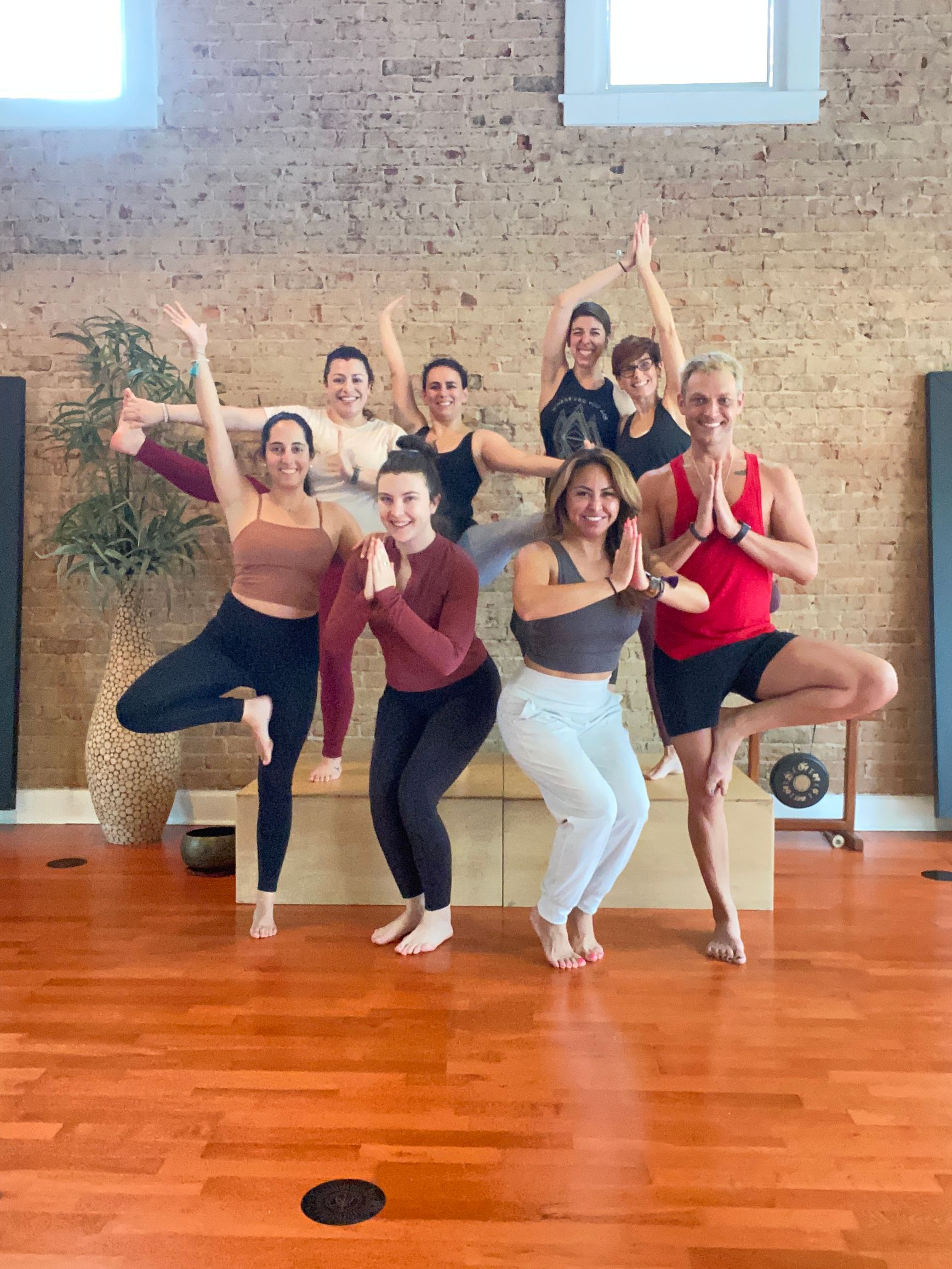 Group of seven people smiling and balancing in tree pose together on wooden blocks inside a brick-walled yoga studio.