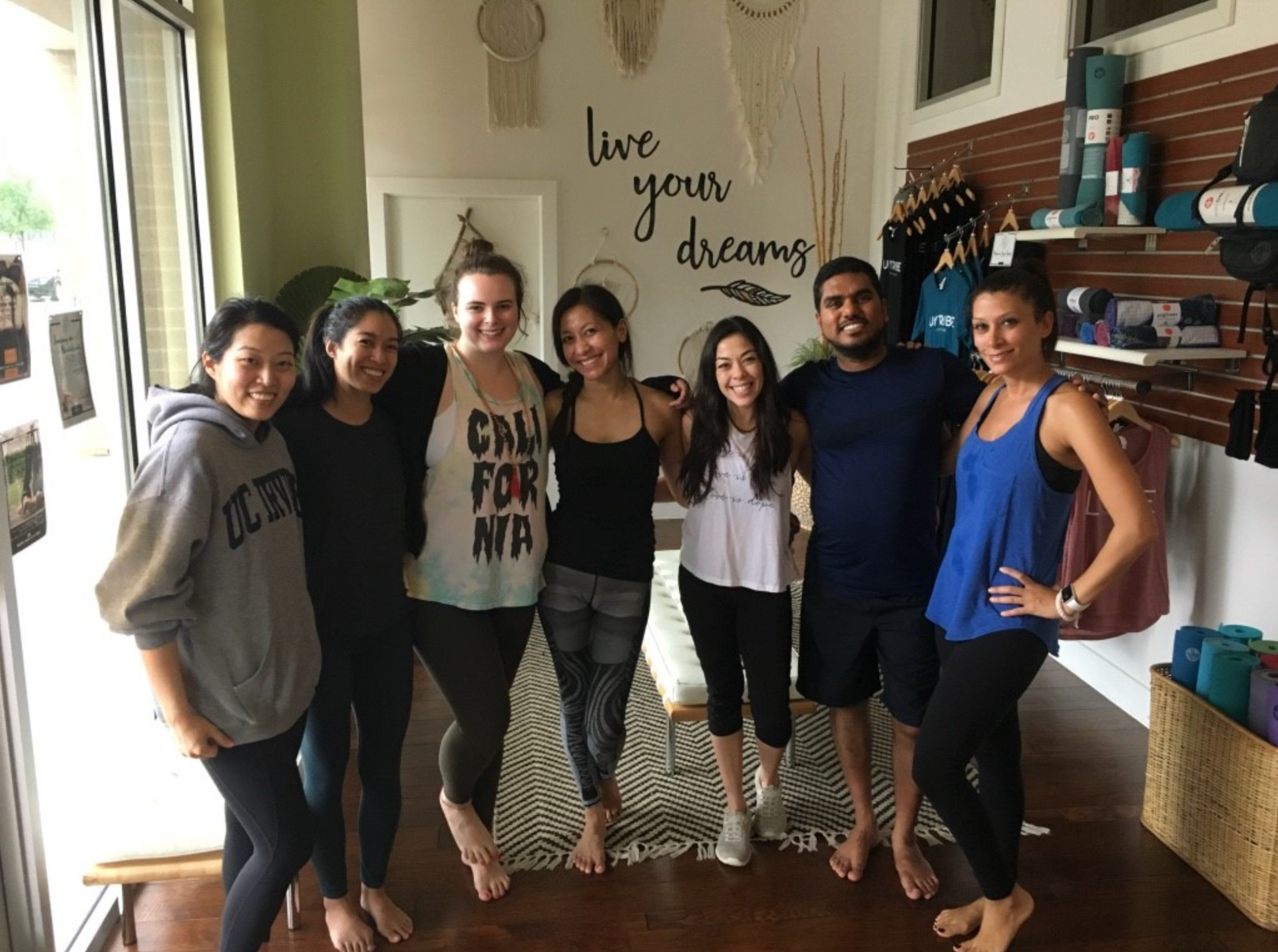 Group of six smiling friends standing in a yoga studio lobby beneath a Live Your Dreams wall decal near retail racks.