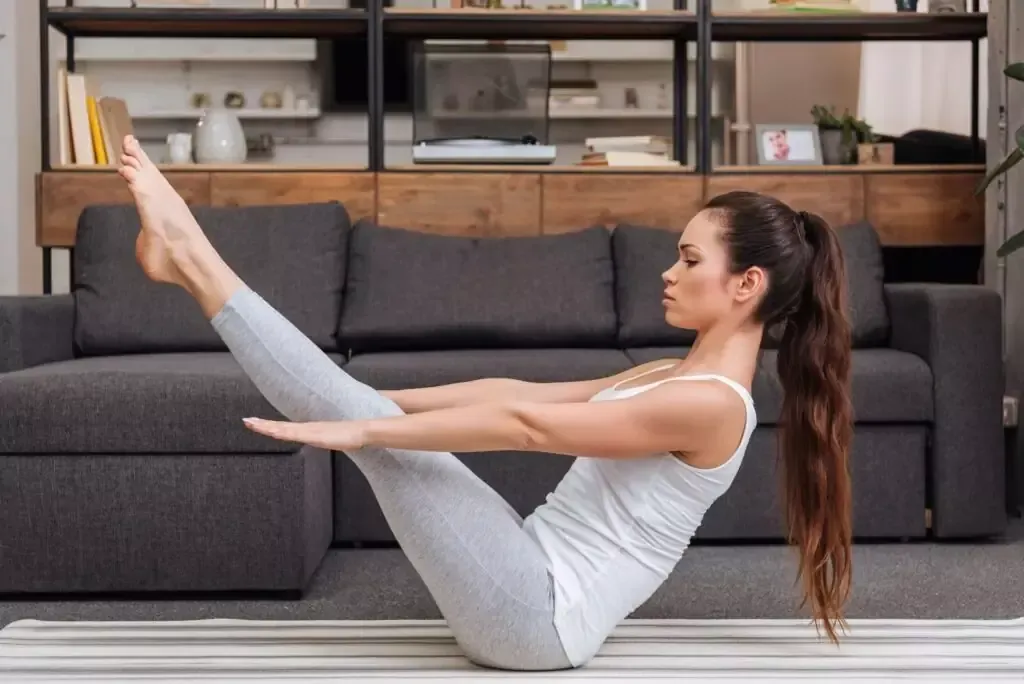 A woman demonstrating Navasana (boat pose) on a yoga mat in her living room, practicing a home yoga routine to build core strength.