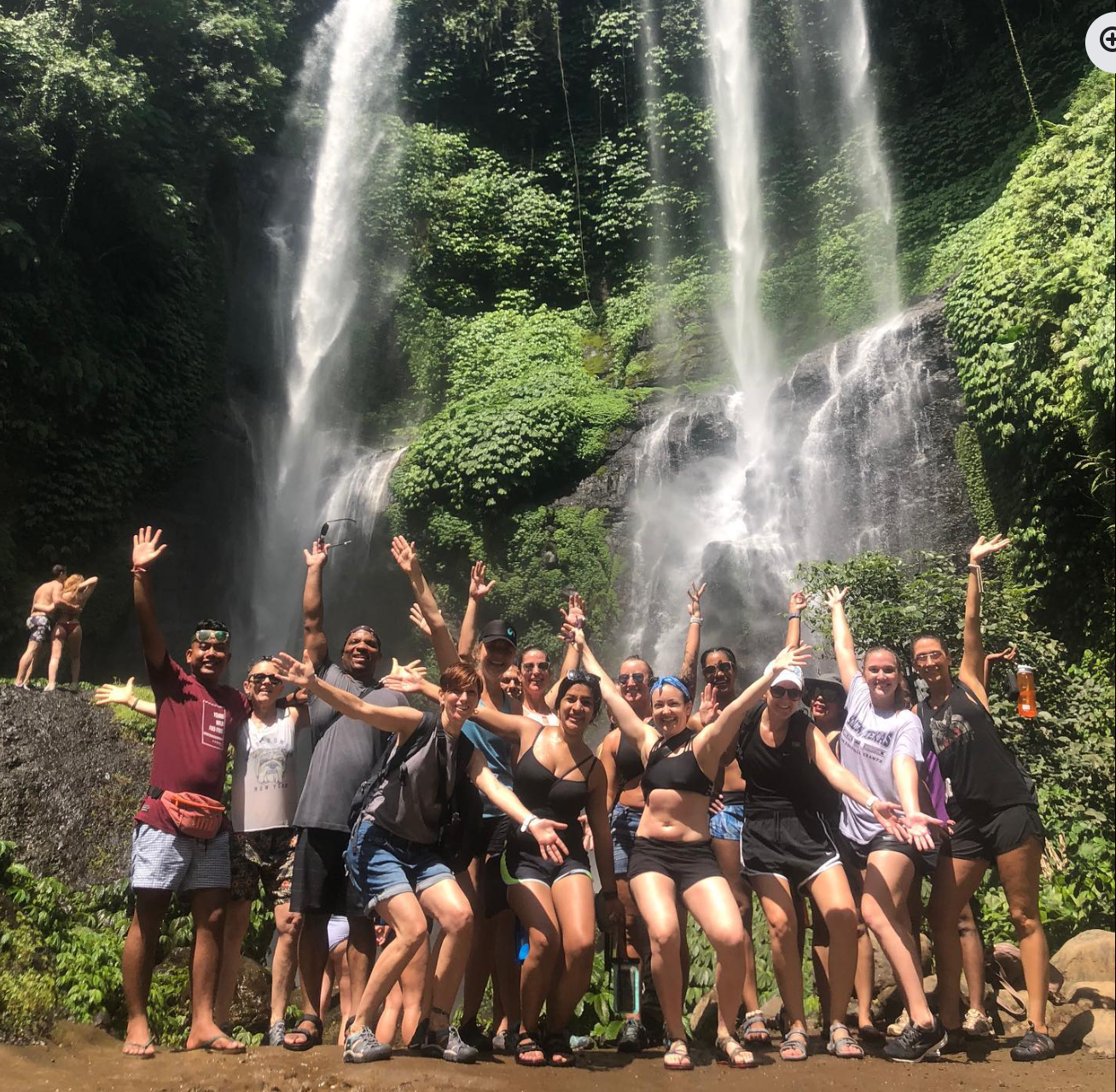 Large group of people cheering with arms raised in front of a massive tropical waterfall during a yoga retreat.