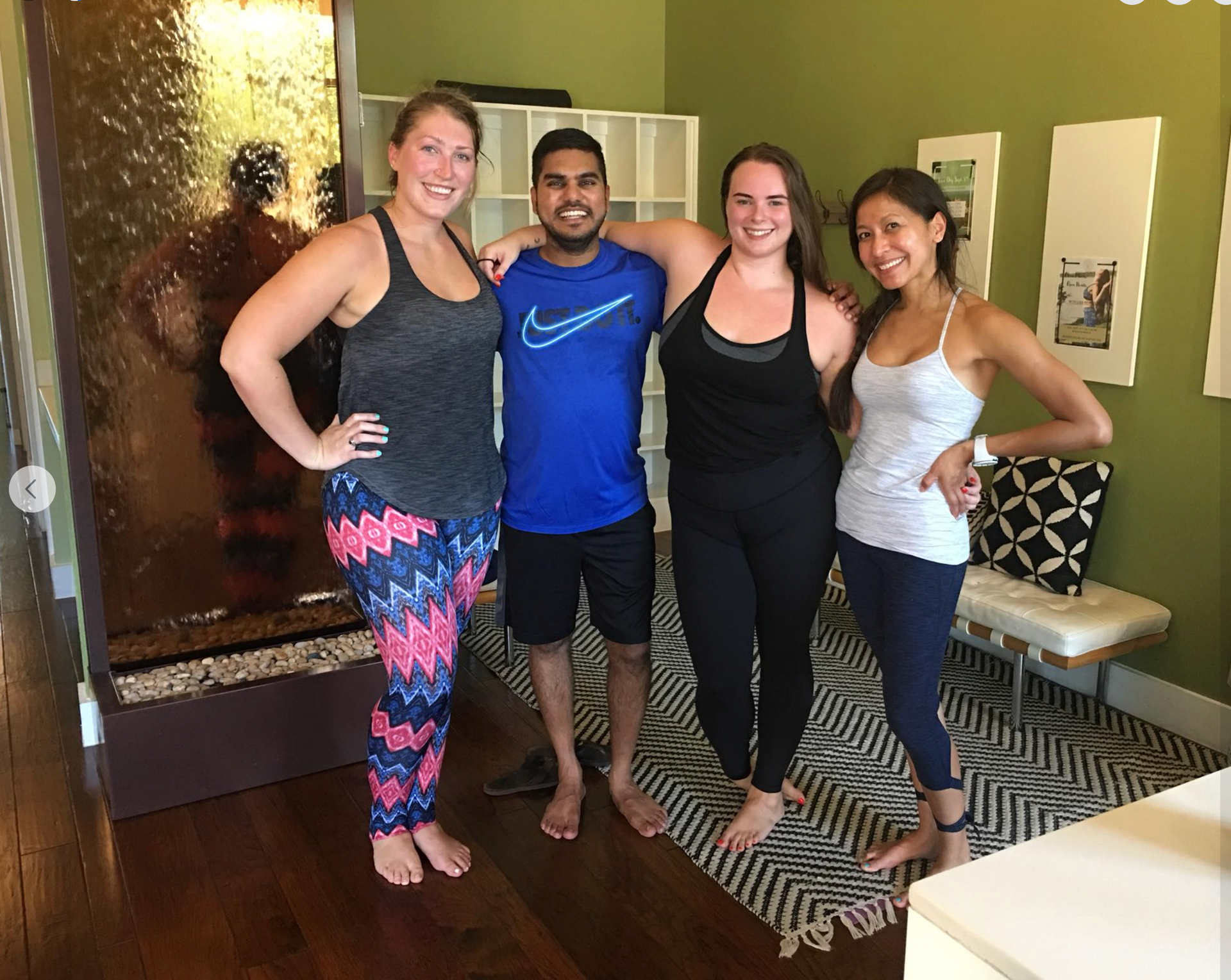 Three women and one man smiling together next to a large indoor waterfall feature in a yoga studio lobby.