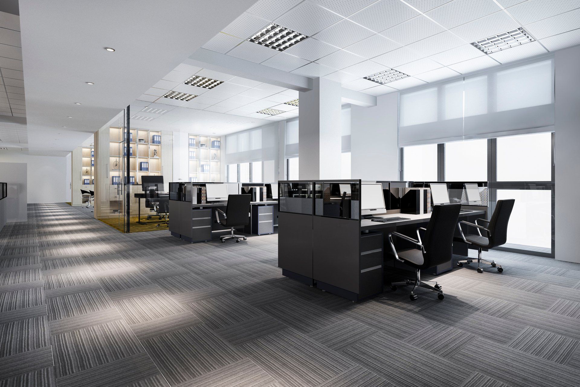 A modern, spacious office featuring rows of dark grey workstations with computers and chairs on a textured grey carpet.