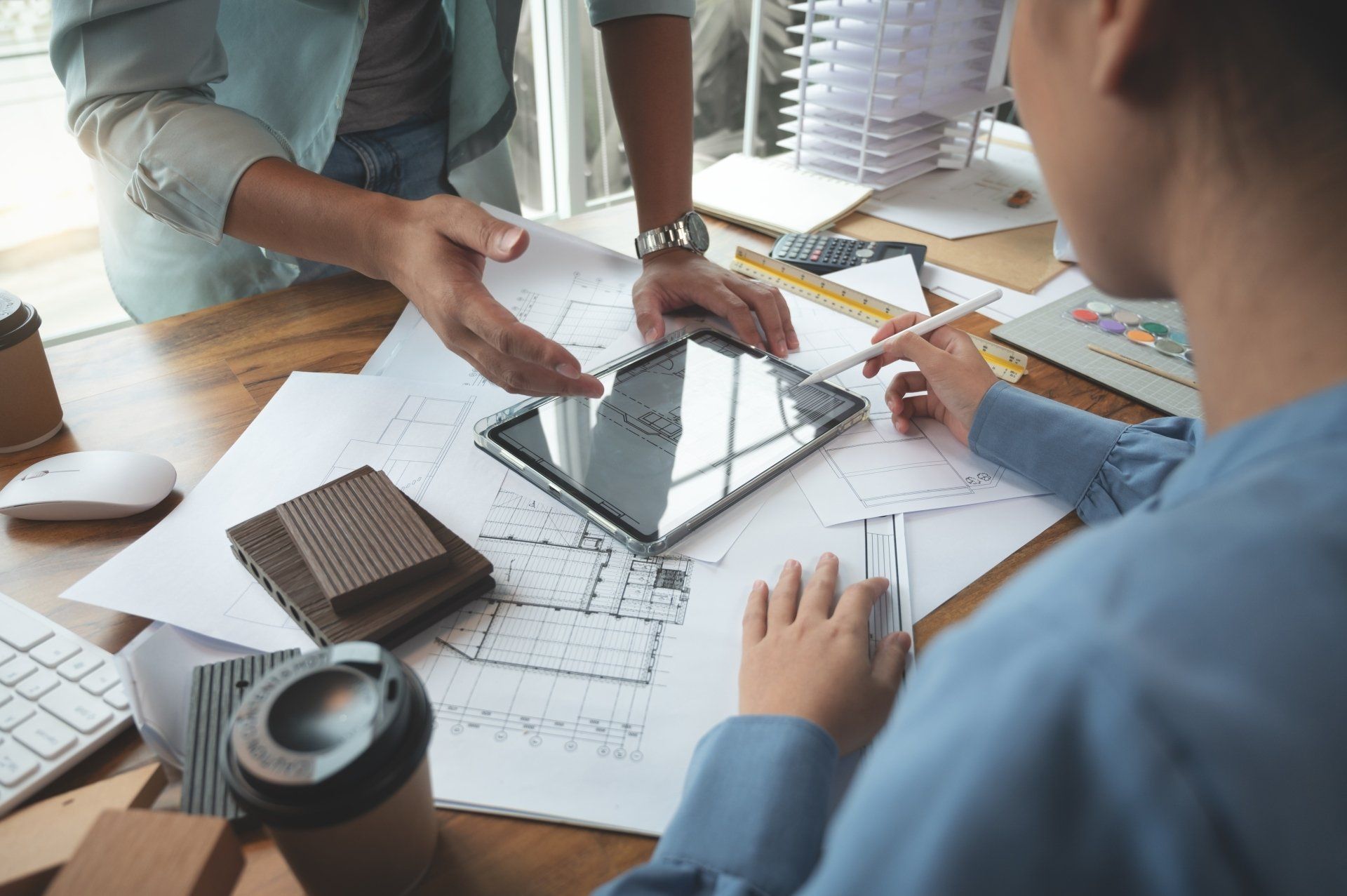 Two people collaborating at a table with blueprints, a tablet, and samples; one points while the other writes.