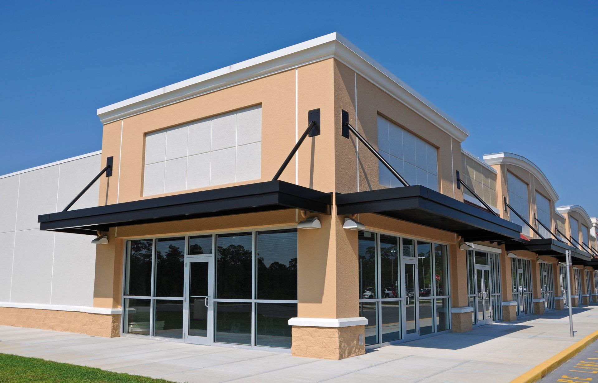 A modern, tan-colored strip mall corner under a clear blue sky, featuring glass storefronts and black metal awnings.