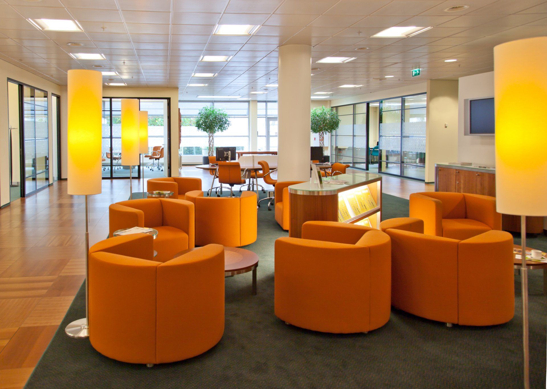 Modern office lounge with orange armchairs, floor lamps, and a central desk on a green carpet, with glass wall partitions.