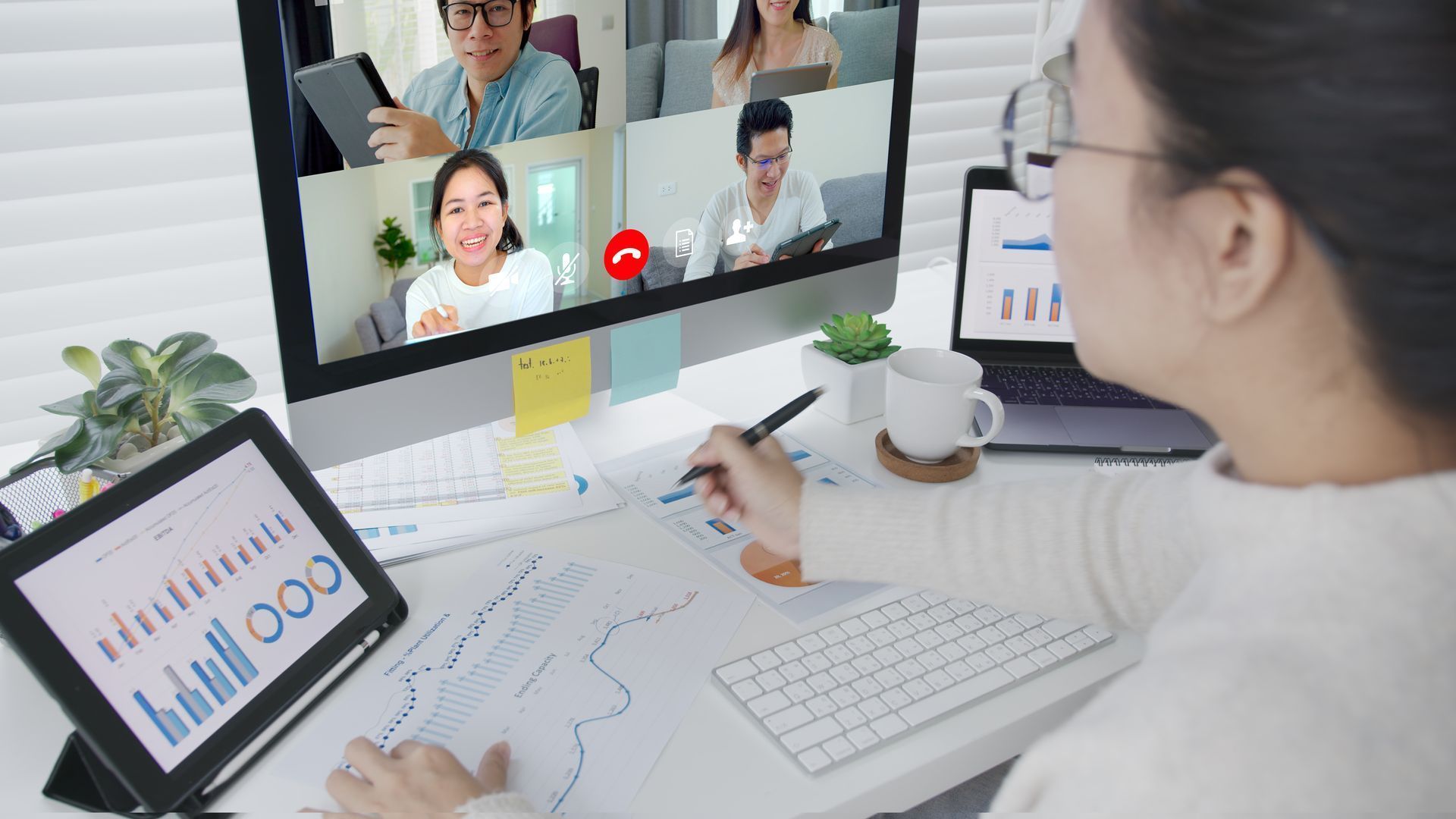 Woman in glasses at desk with video call on monitor, looking at graphs on tablet and paper.