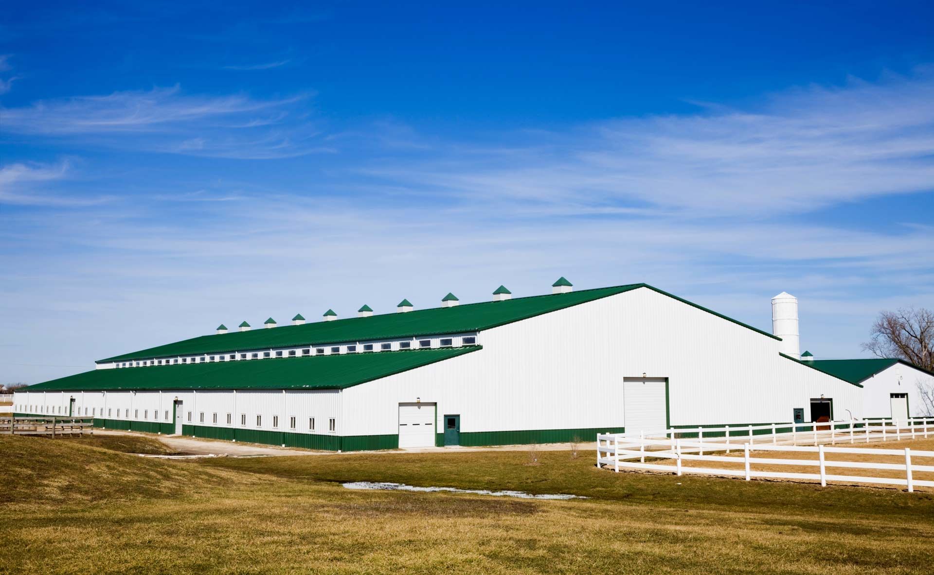 White and green barn with a green roof against a blue sky, on a grassy field with a white fence.