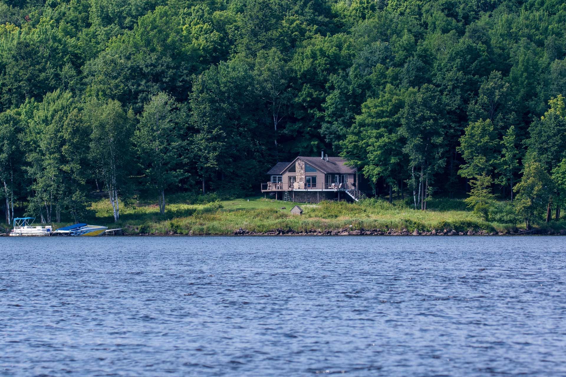 A house on a lake, surrounded by green trees. Boats are visible on the left.