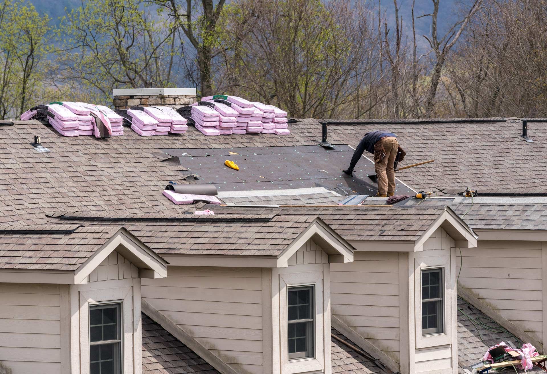 Roofer working on a residential roof, surrounded by supplies. Light brown shingles, bright pink insulation. Overcast sky.