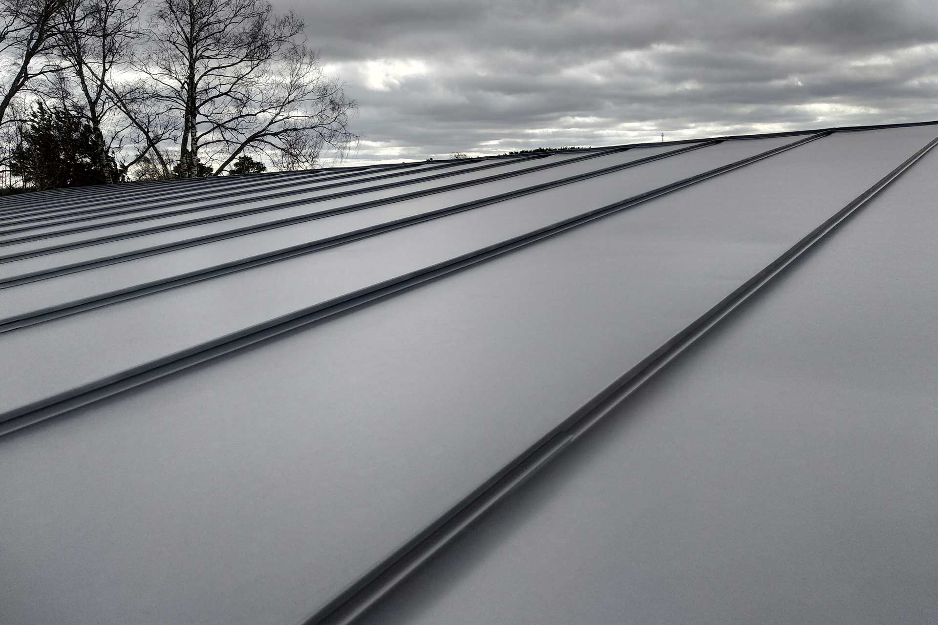 Close-up of a silver metal roof with ribbed panels and a cloudy sky background.