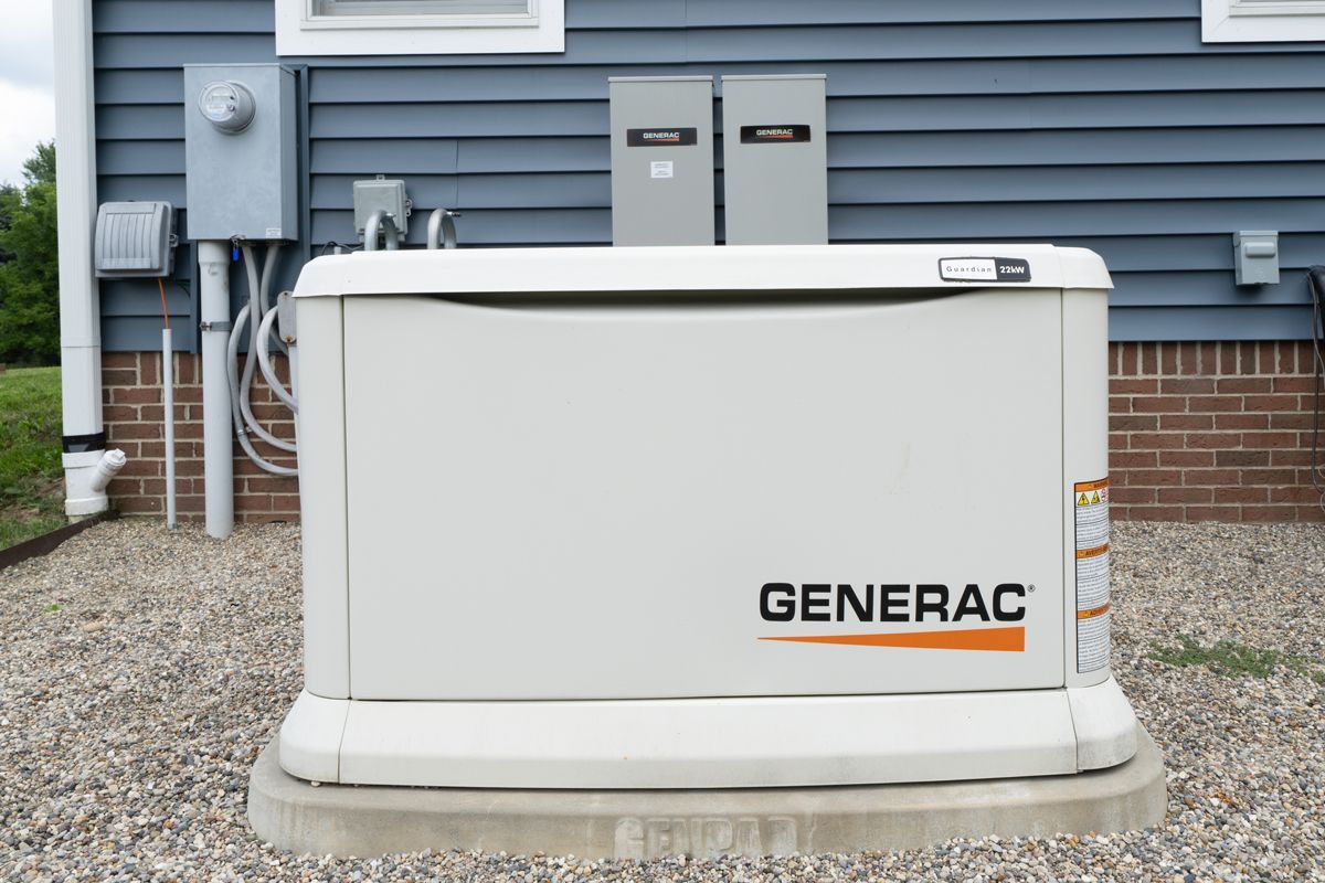 Generac home generator on a gravel bed, installed near a blue-sided house with electrical components.