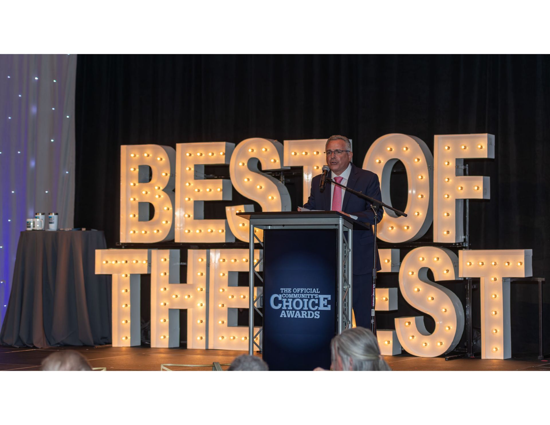 A person in a suit speaks from a podium in front of large, illuminated letters reading