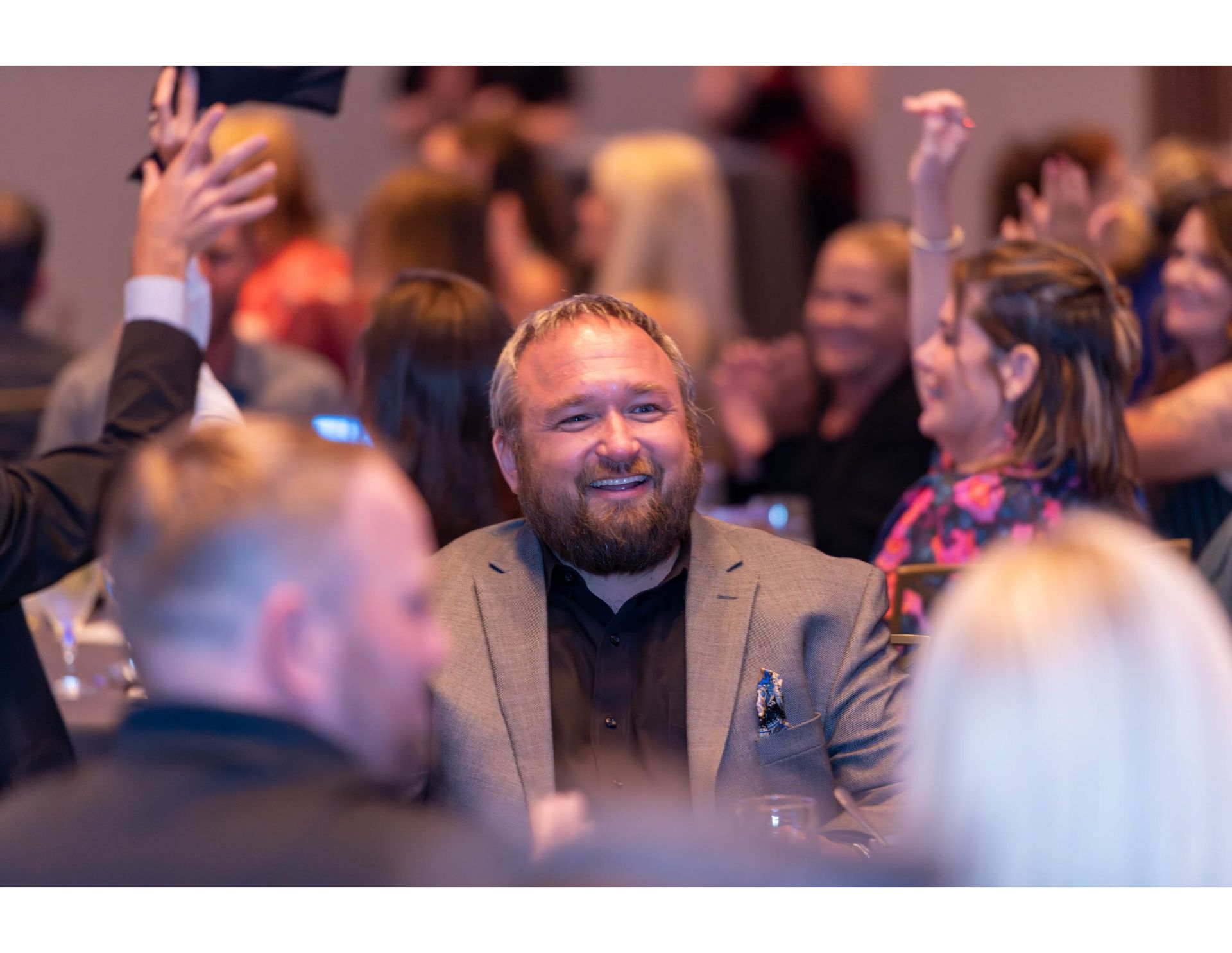 A smiling person in a suit sits at a table in a crowded, blurred event space while others raise their hands in the air.
