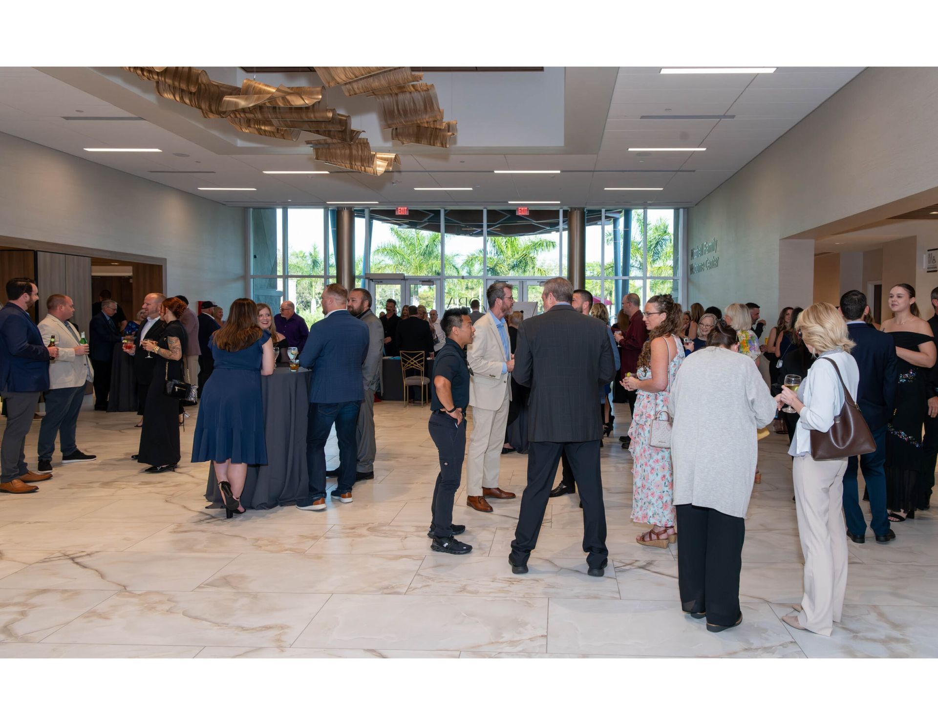 A social gathering with people in business-casual attire mingling in a bright, spacious hall with marble flooring.