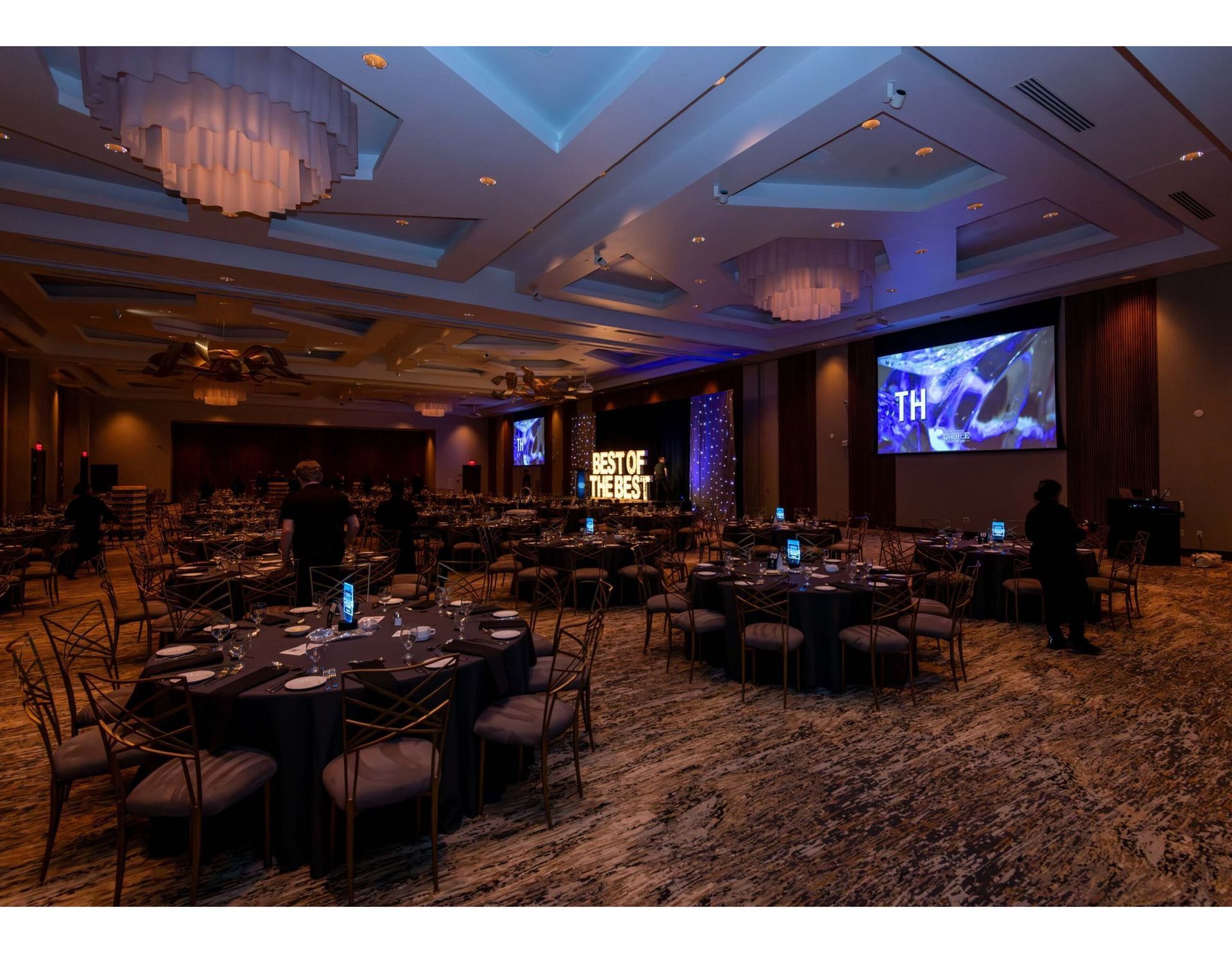 A large hotel banquet hall set up for an event, featuring round tables with black tablecloths and large projection screens.