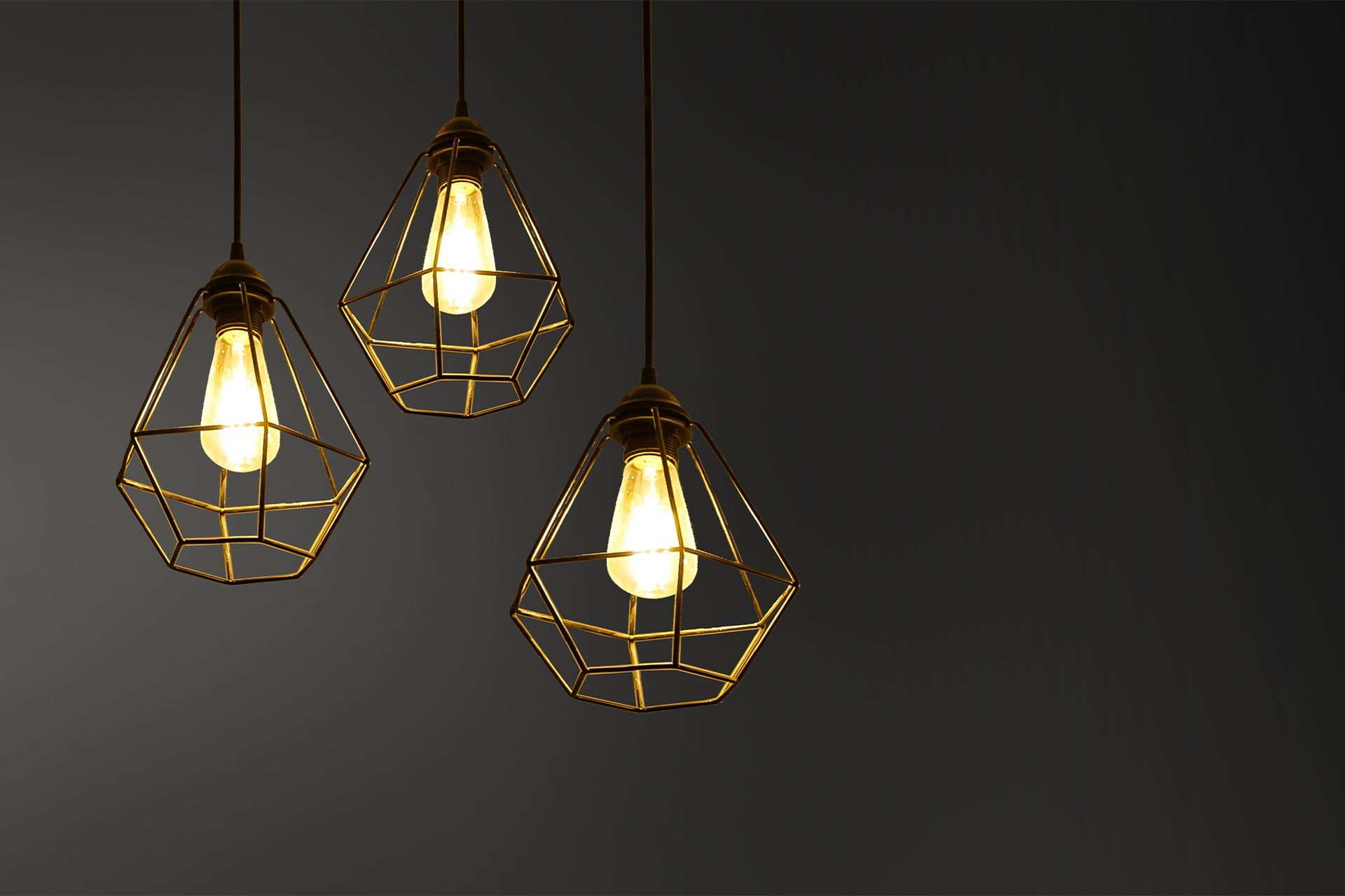 Three pendant lights with gold geometric cages, lit bulbs against a dark gray background.