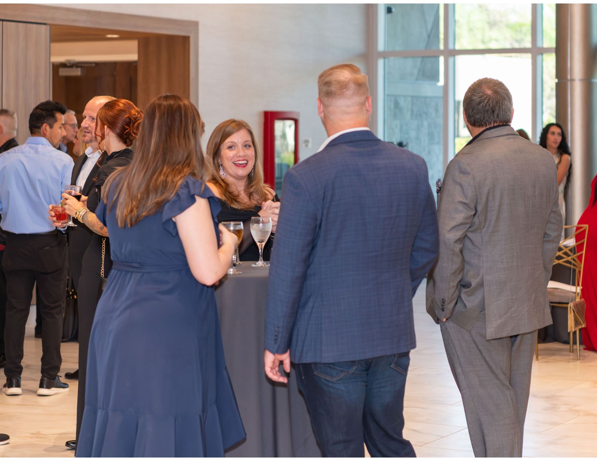 A group of people in business-casual attire chatting and holding drinks while standing at a reception in a bright lobby.