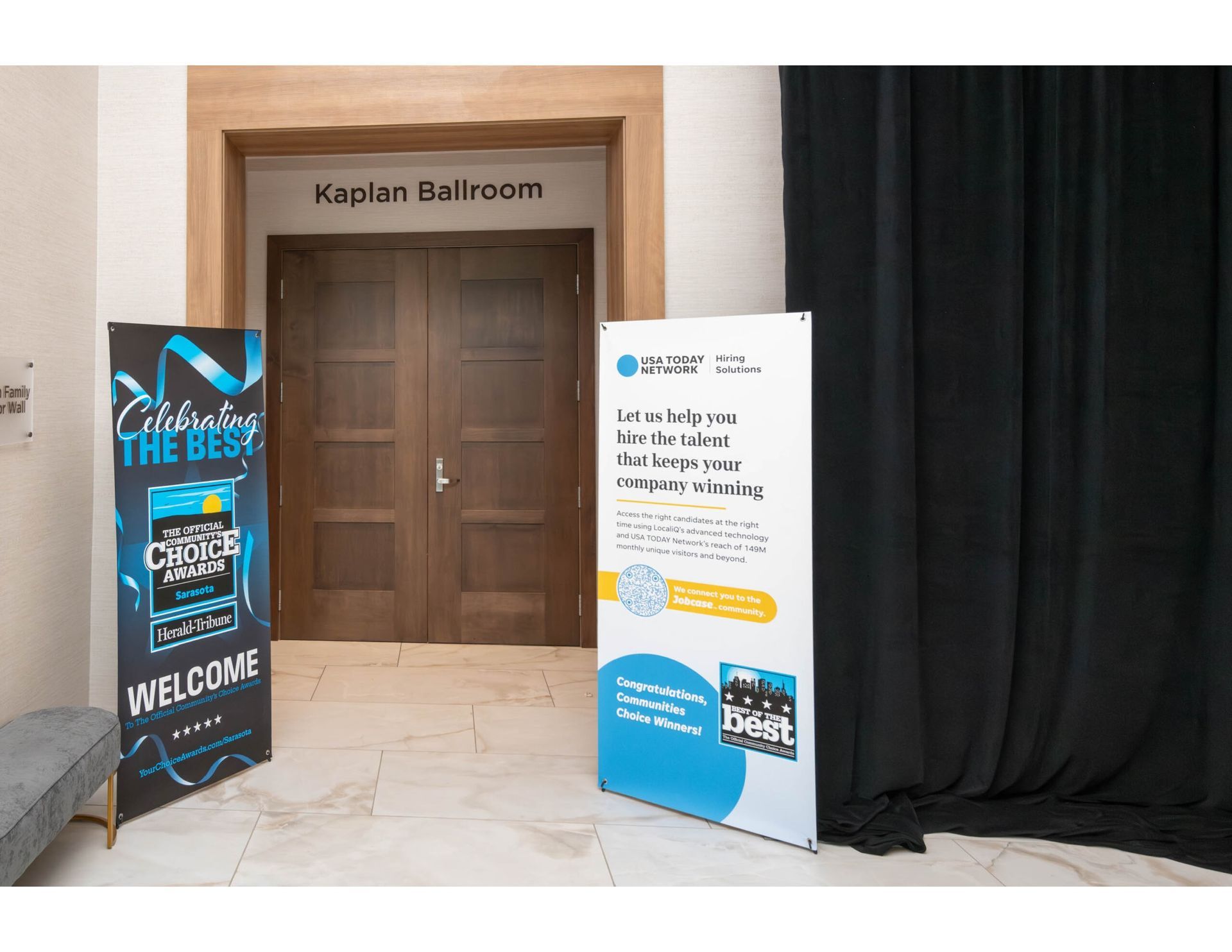 Two vertical event banners stand outside the Kaplan Ballroom entrance, which has double wooden doors and black curtains.