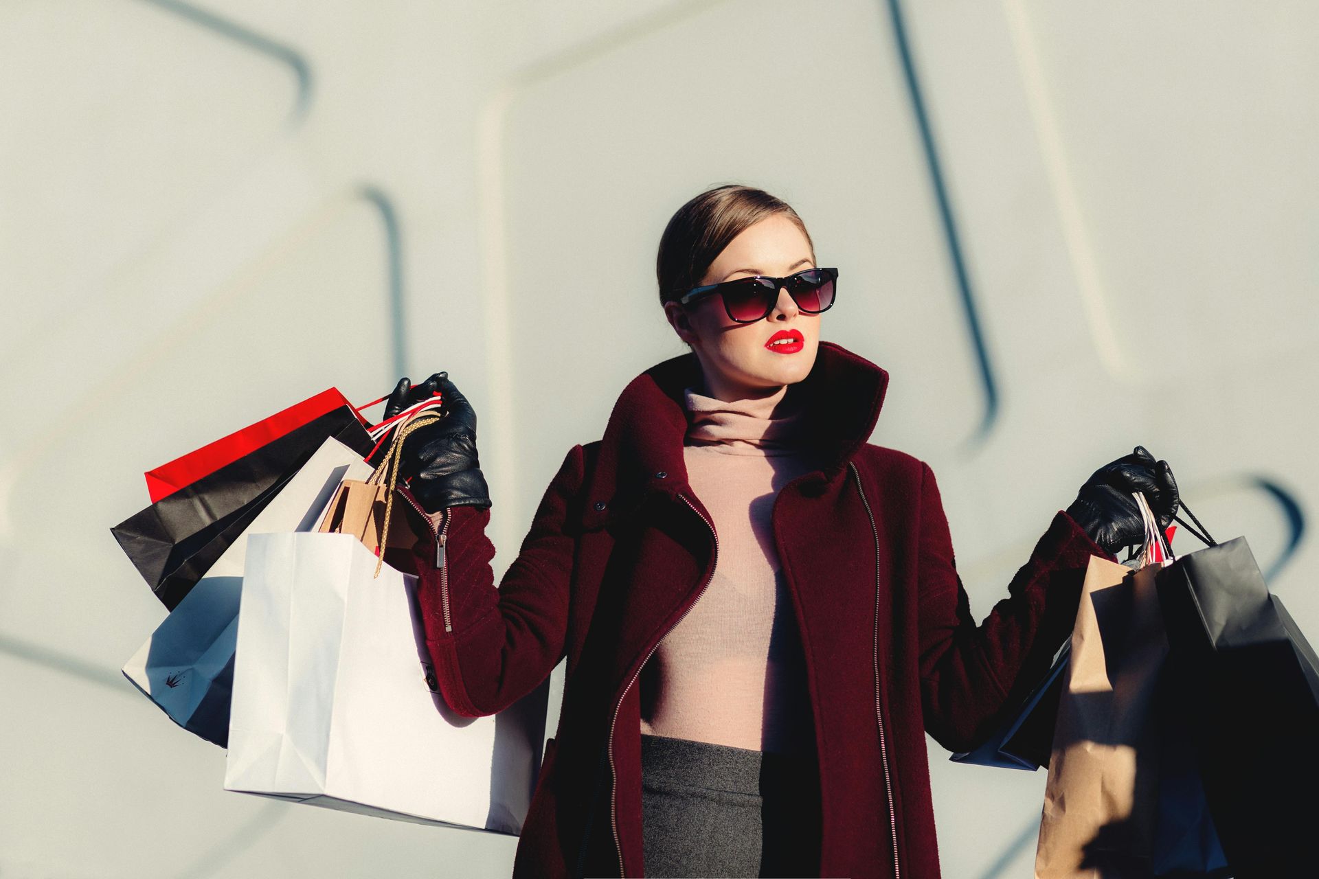 Donna con cappotto rosso e occhiali da sole che tiene in mano delle borse della spesa all'esterno.
