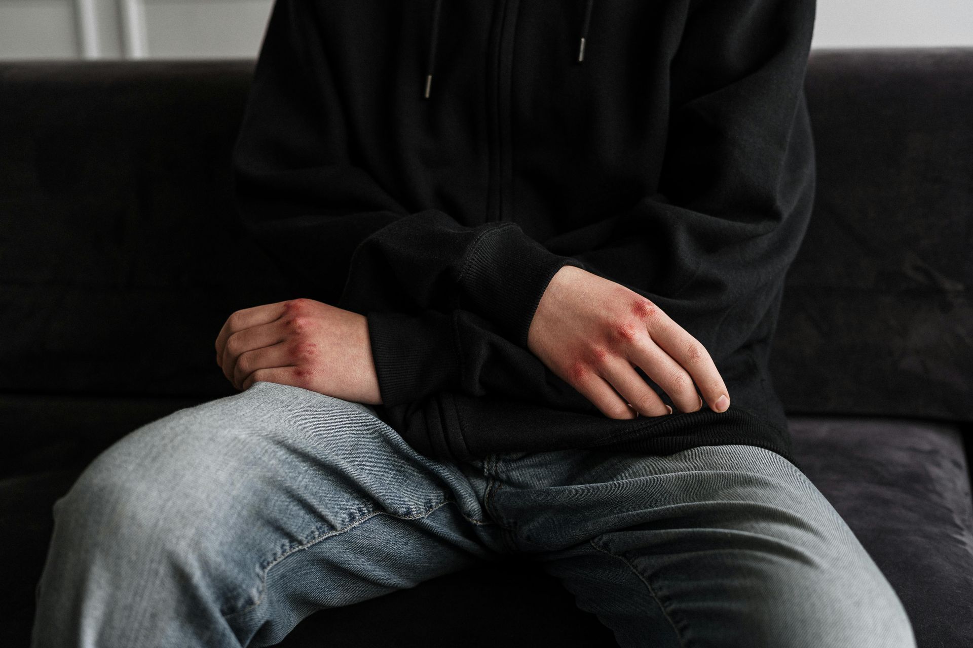Persona con felpa nera e jeans seduta su un divano nero, con le mani in grembo.