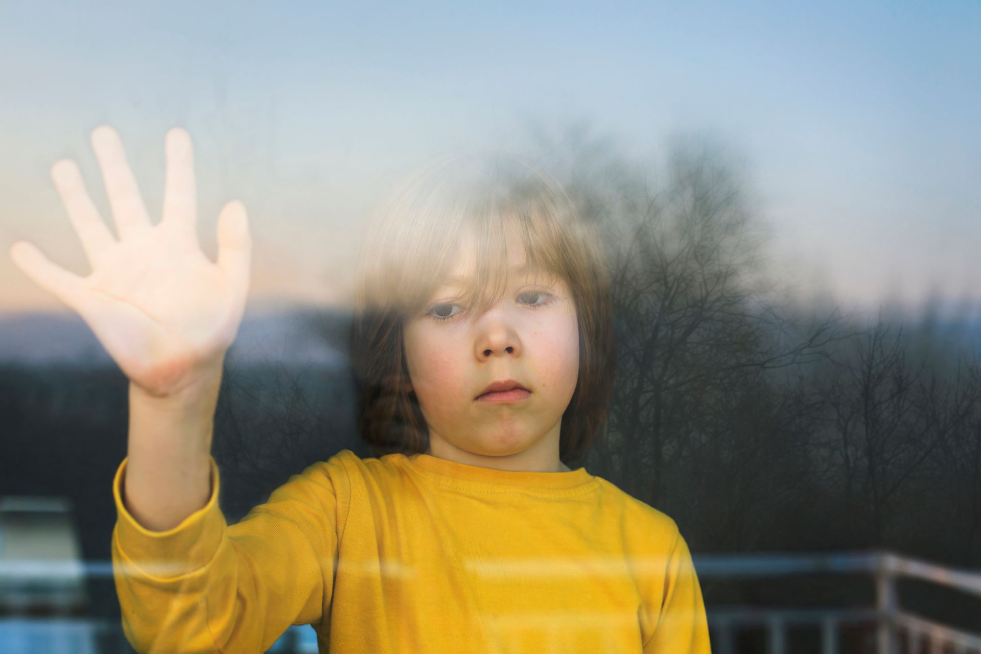 Ragazzo con la mano appoggiata alla finestra, dall'aria triste; all'esterno, cielo coperto.