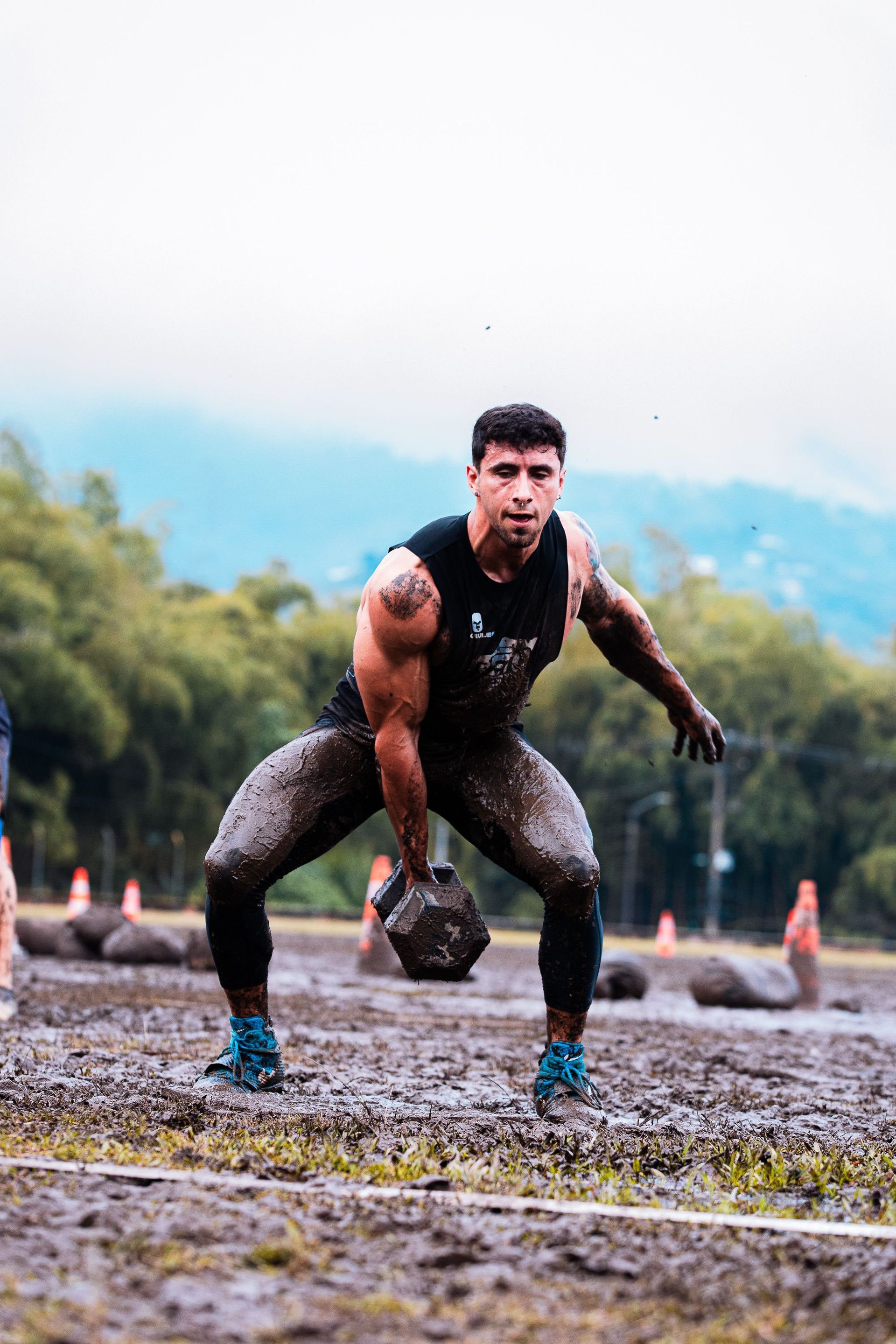 Athlete in black gear lifting a kettlebell on a muddy outdoor track, with hills in the background