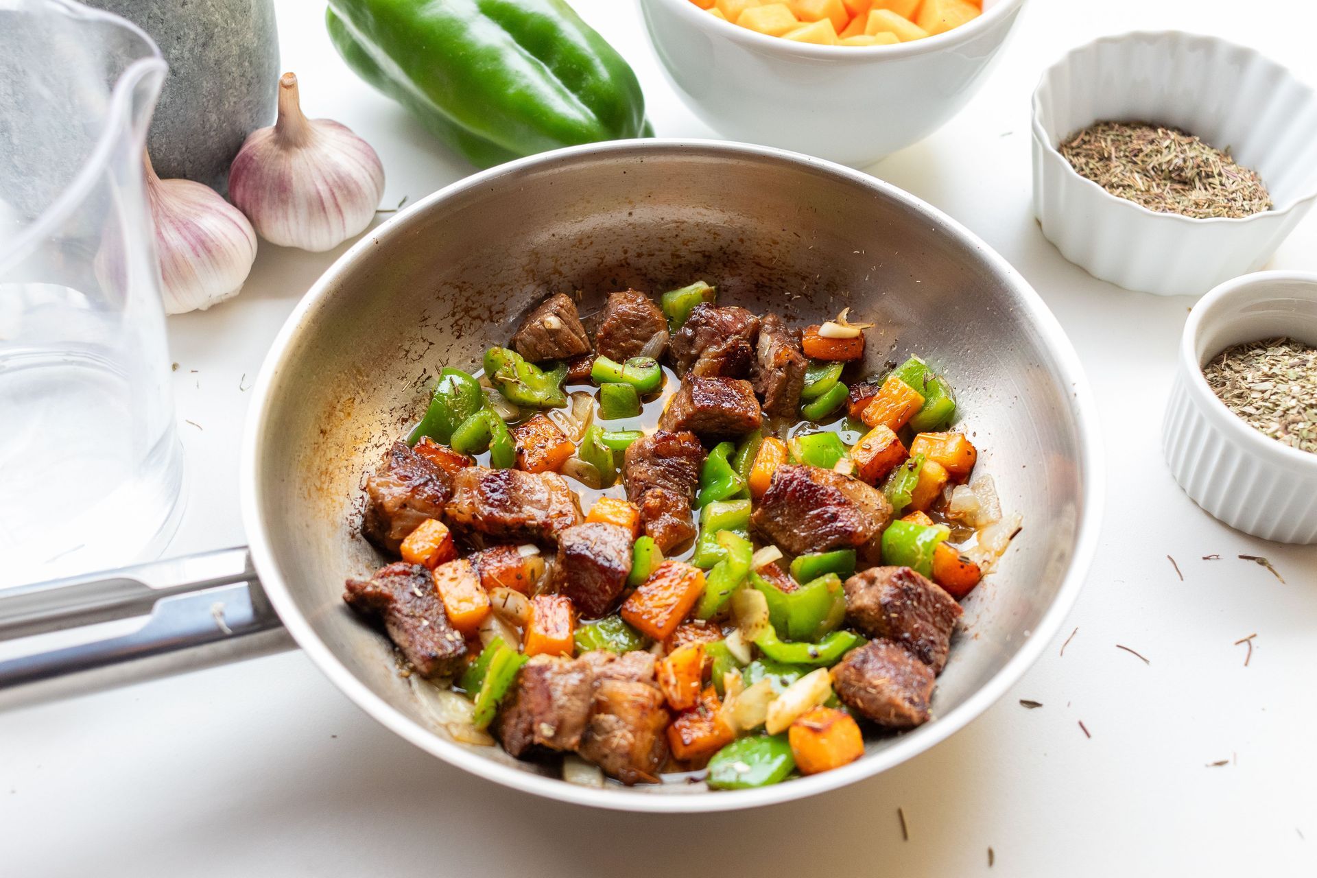Pan of sautéed diced meat and green bell peppers on a white countertop with spices nearby